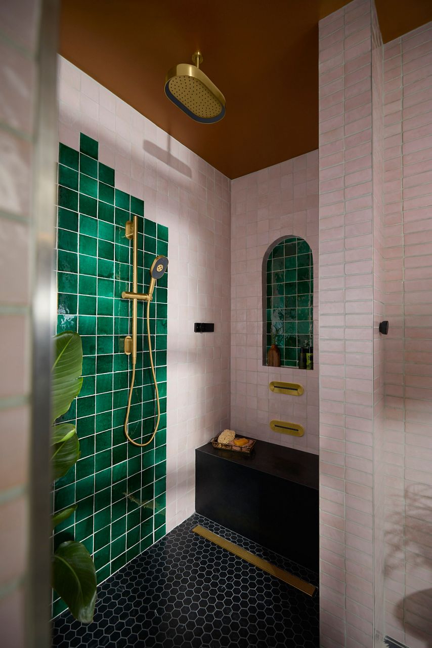 A vibrant shower with lustrous deep green tiles, complemented by gold fixtures, including a rain shower head and handheld wand. A built-in black shelf and a small arched inset provide space for toiletries. The shower radiates a lush, opulent feel with a contrasting dark floor.