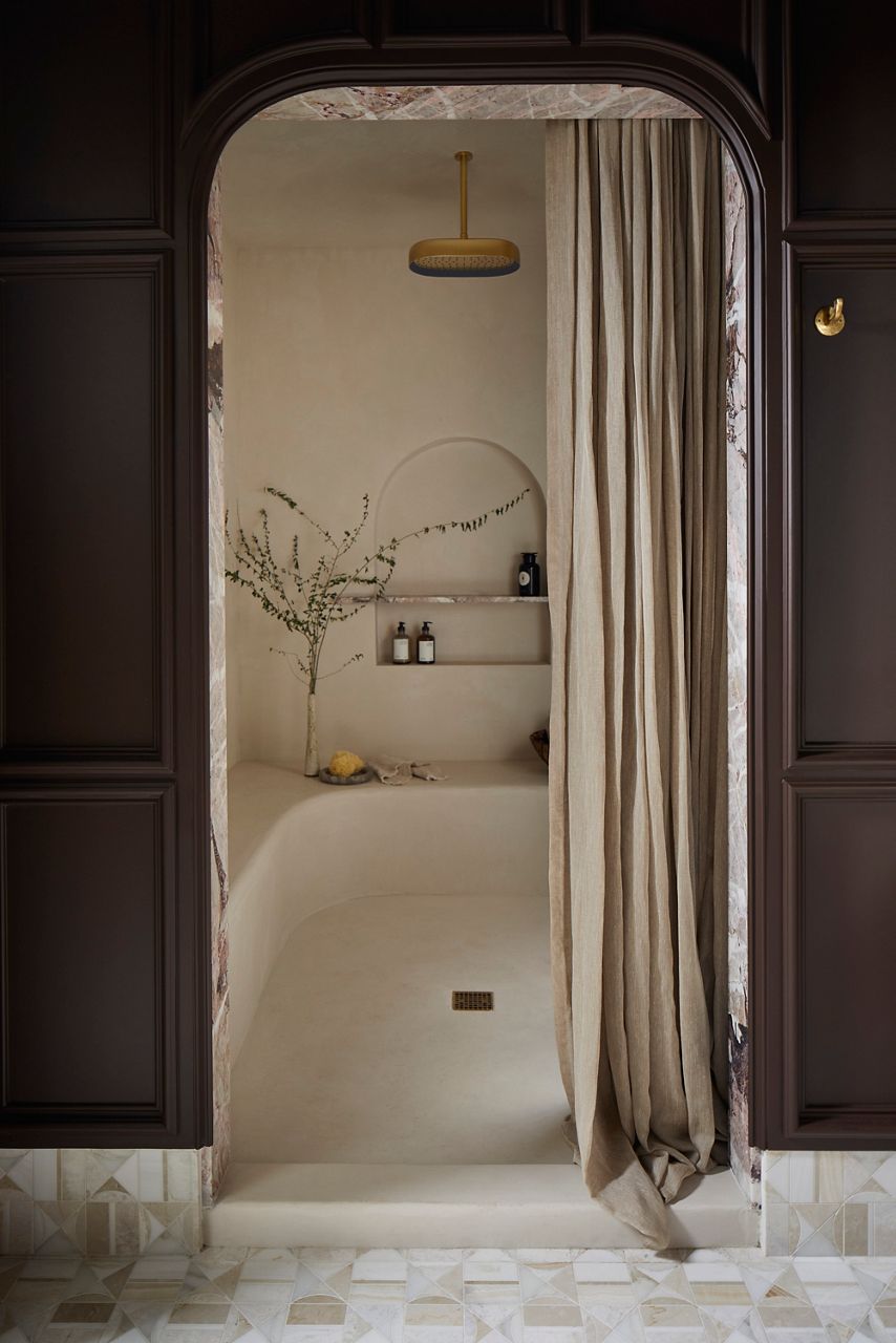 An arched alcove shower elegantly framed by dark wood panels, with a flowing white curtain pulled slightly aside. The interior features a built-in bench and recessed shelves holding bath essentials, all under a brass rain shower head, with soft lighting creating a tranquil, spa-like atmosphere.