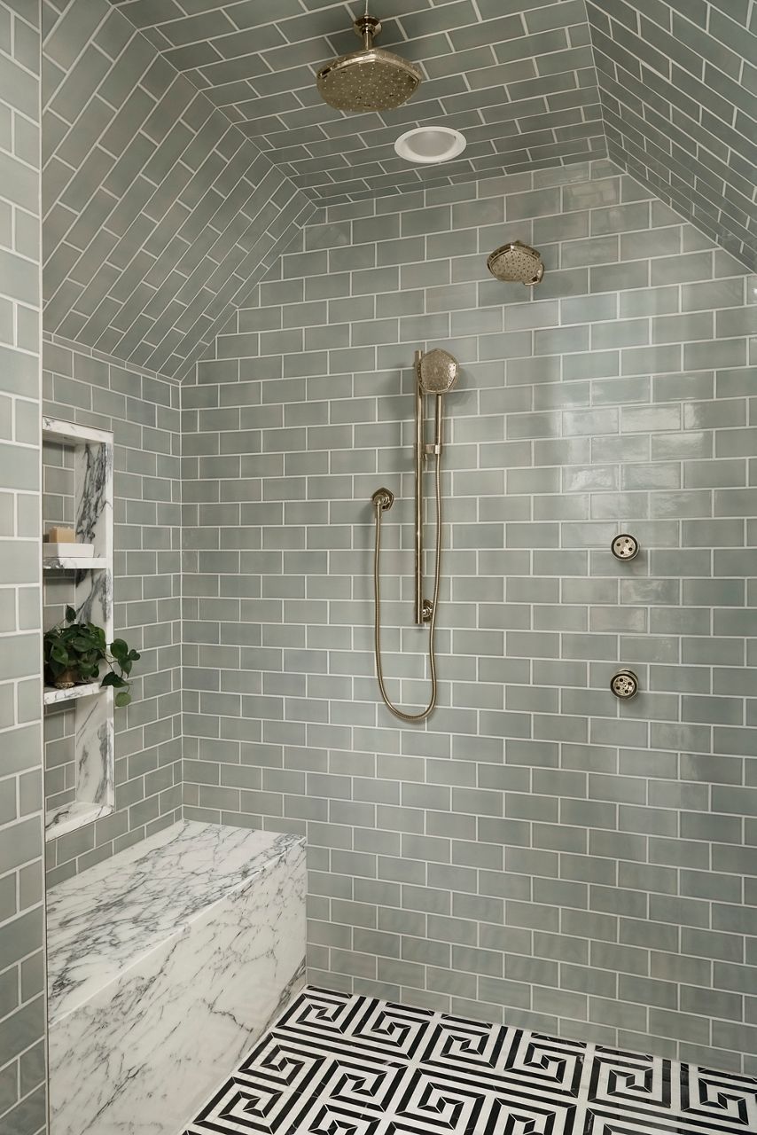 Shower alcove with sage green subway tiles and marble flooring. It features a gold rain shower head, handheld spray, and three body jets. A built-in niche for toiletries and a small bench add practicality and charm.
