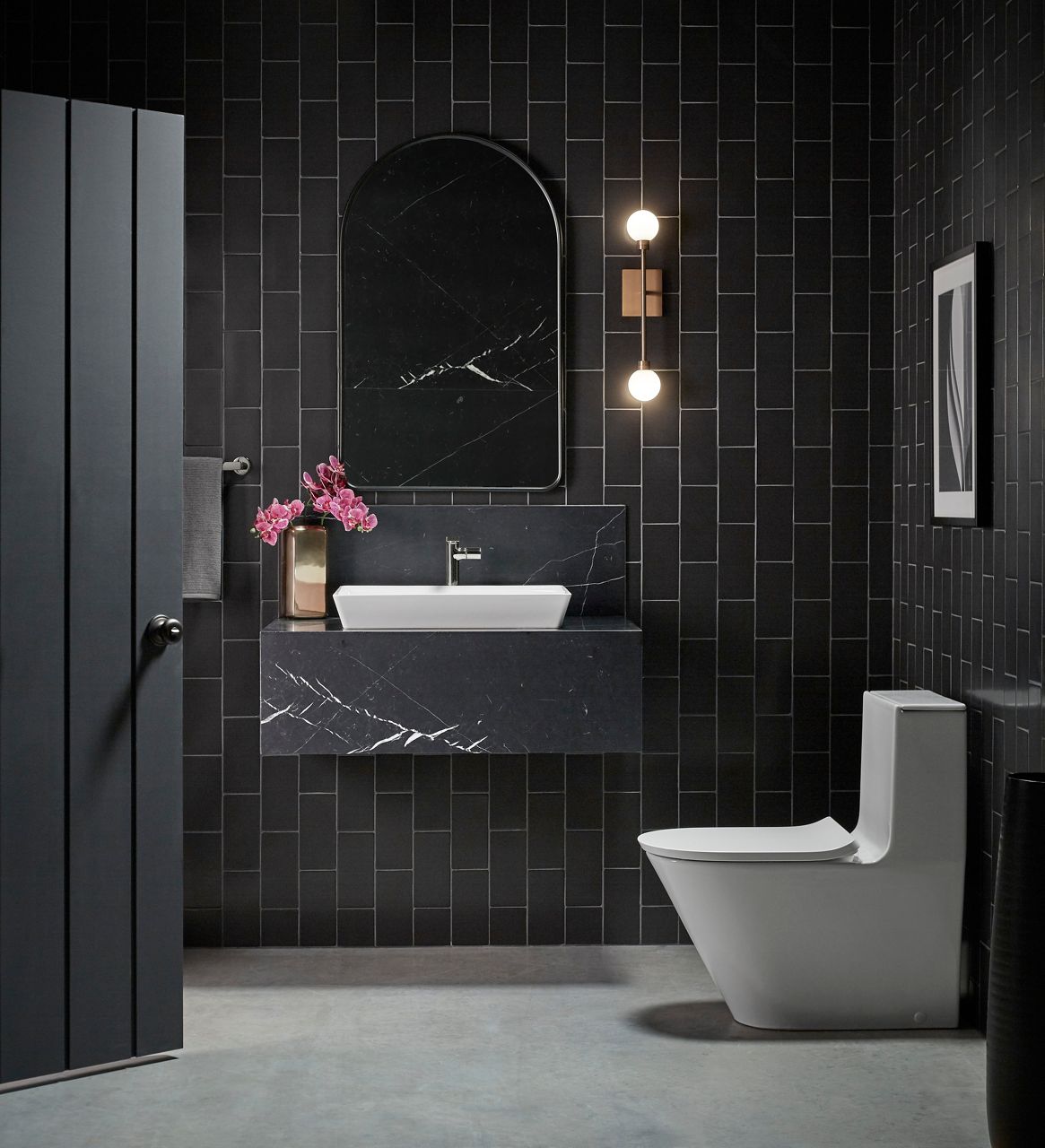 A white Brazn sink and toilet in a modern bathroom with vertically-oriented rectangular black tile with light gray grout, a black quartz wall-hung vanity and dark pink orchids.