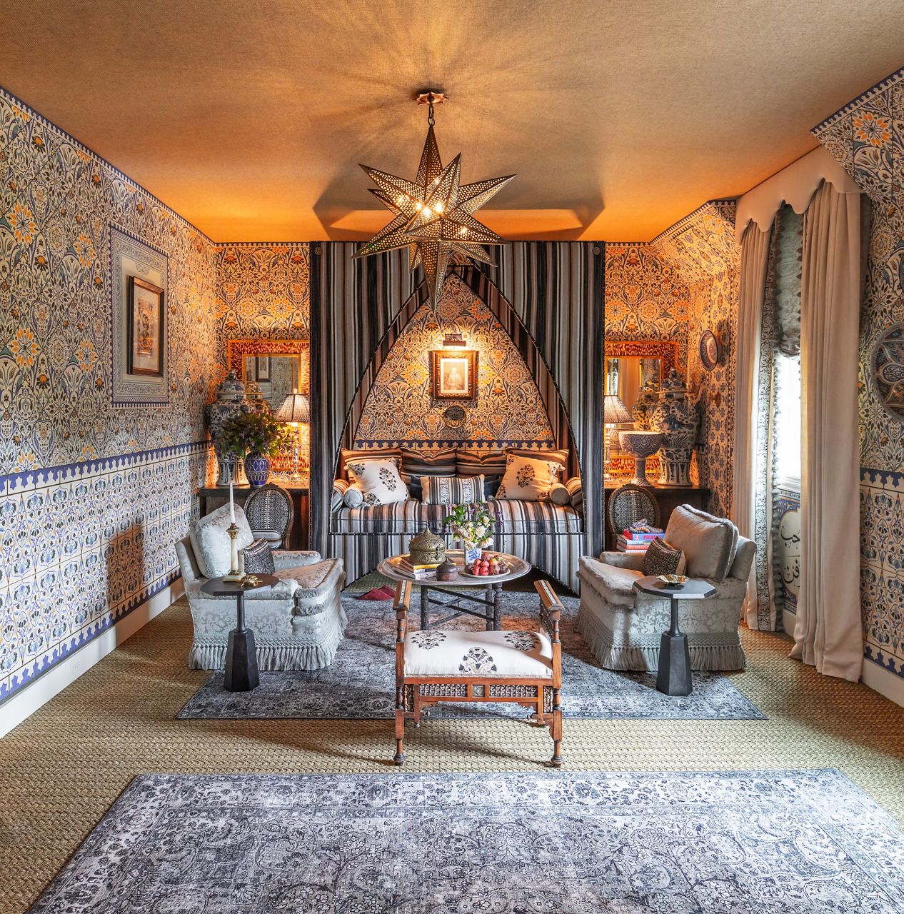Un salon de style grand-millénaire présente un papier peint fleuri, un lit de jour rayé avec baldaquin, des pots de gingembre bleus et blancs, et des fauteuils à imprimé floral se faisant face au-dessus d’une table basse ronde.