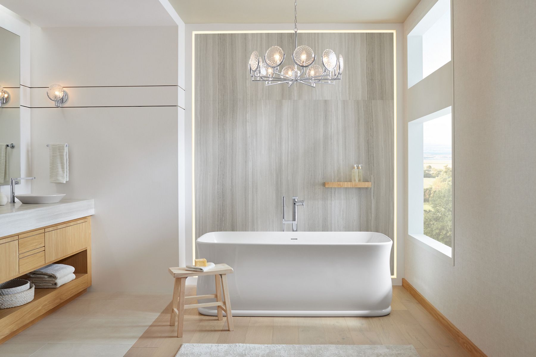 Daylight in modern bathroom with freestanding bath and illuminated wall sconces and chandelier.