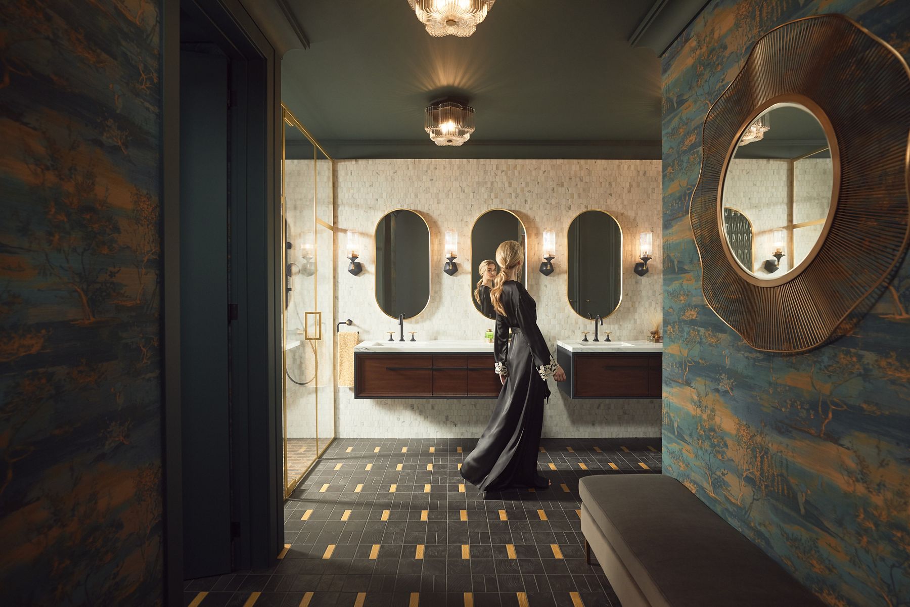 Une femme blonde vêtue d’une longue robe noire regarde sa réflexion dans une salle de bain avec deux meubles-lavabos Lodern de KOHLER à installation murale, des robinets à poignée à levier déployé Occasion et trois miroirs Essential en forme de capsule.