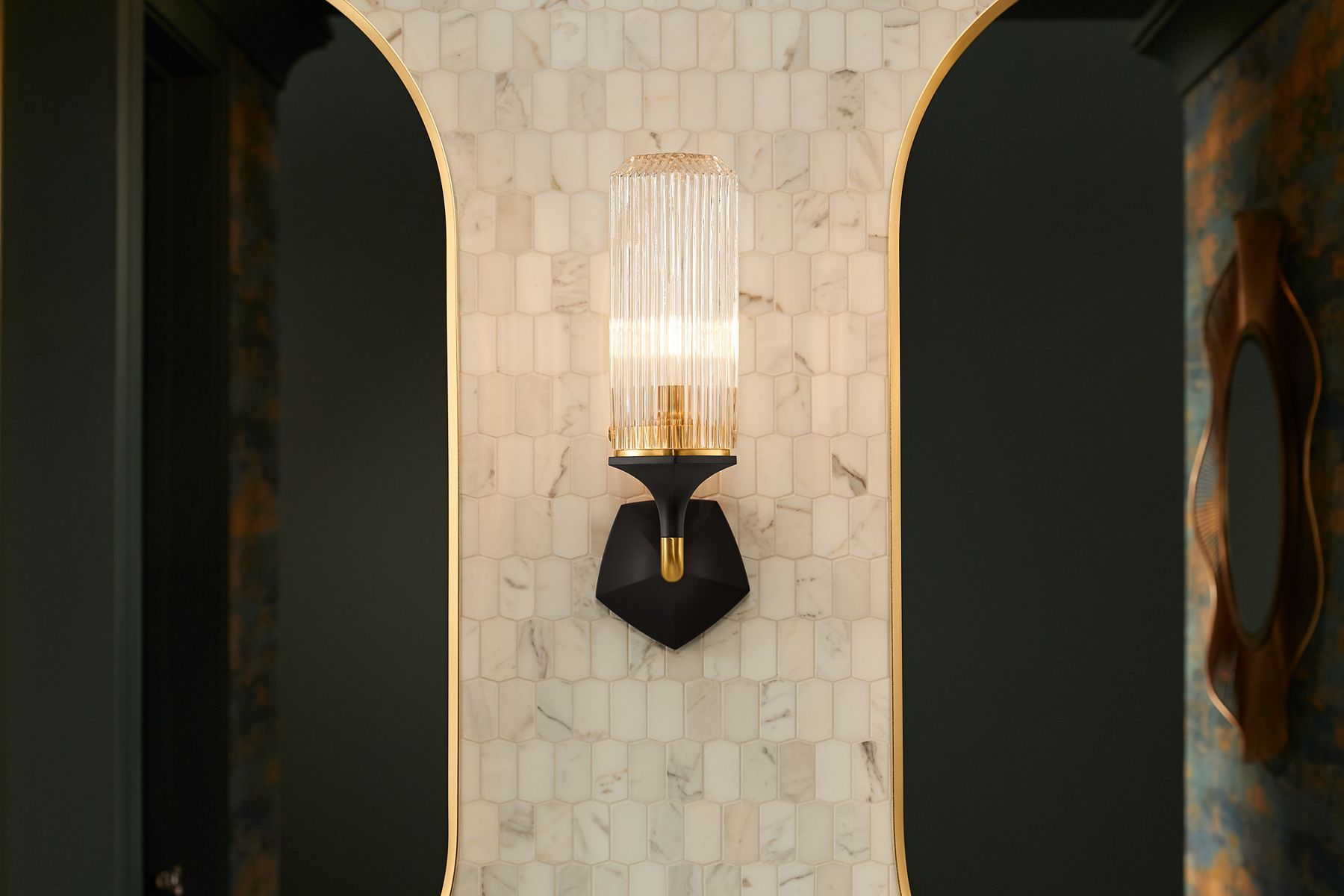 An Occasion wall sconce in Matte Black and Brushed Moderne Brass two-tone finish against a wall of small cream-colored marble tiles.