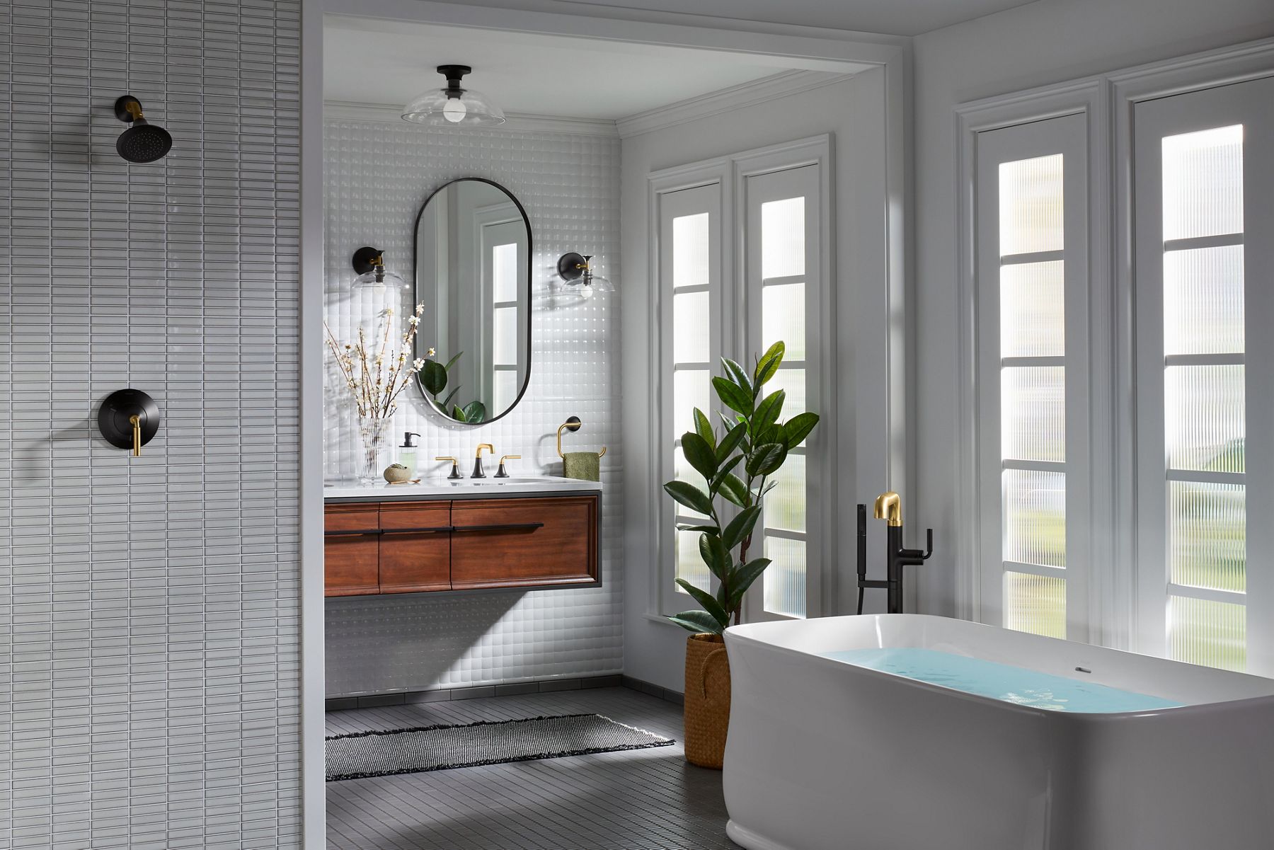 Une salle de bain avec douche à carreaux gris, meuble-lavabo mural Lodern de KOHLER, miroir en forme de capsule et baignoire autoportante Imperator avec remplisseur de baignoire autoportante Tone.
