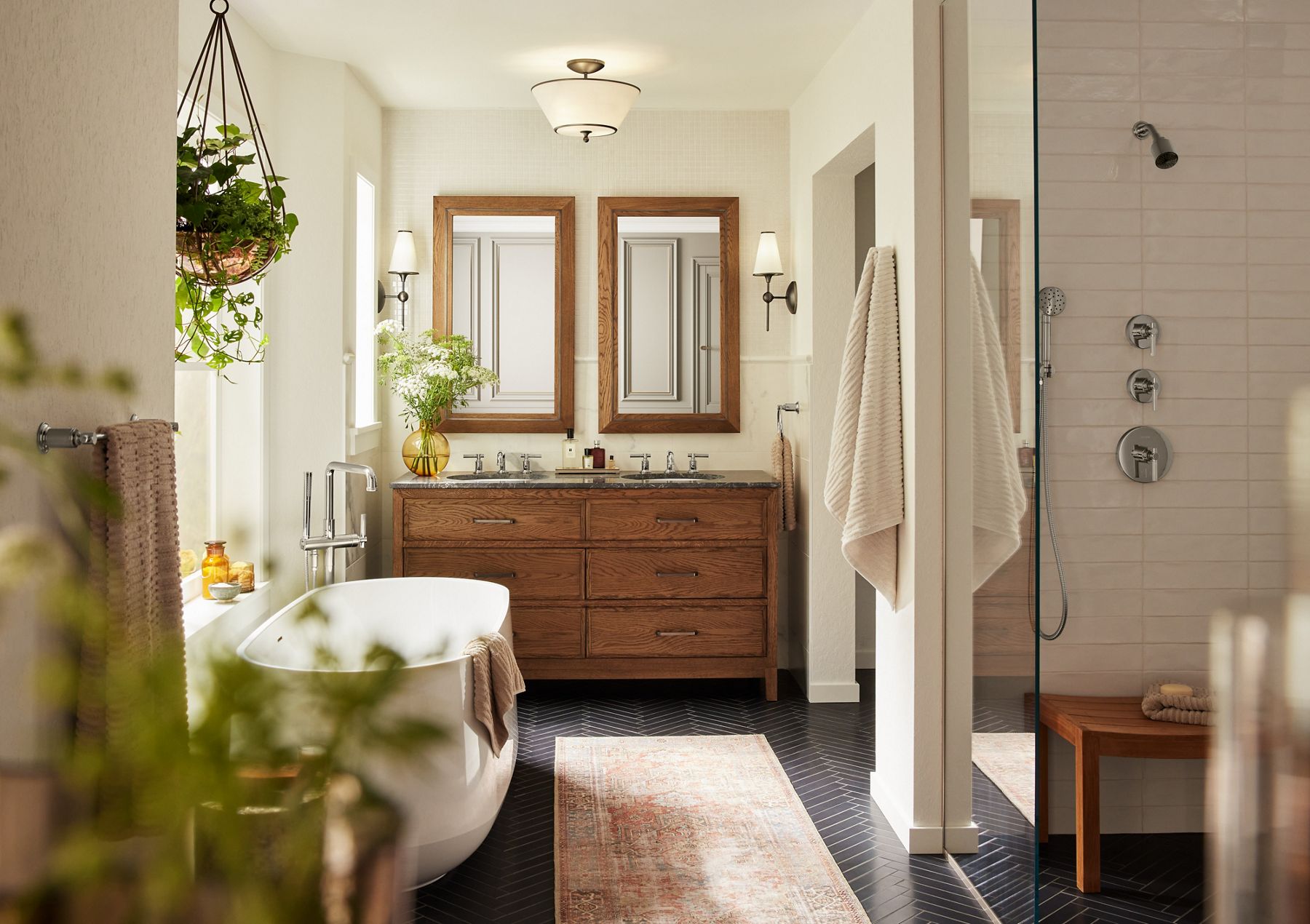 A sunfilled bathroom with a freestanding bath on the left, shower area on the right, wood grain double vanity in the back, and dark gray herringbone patterned floors.