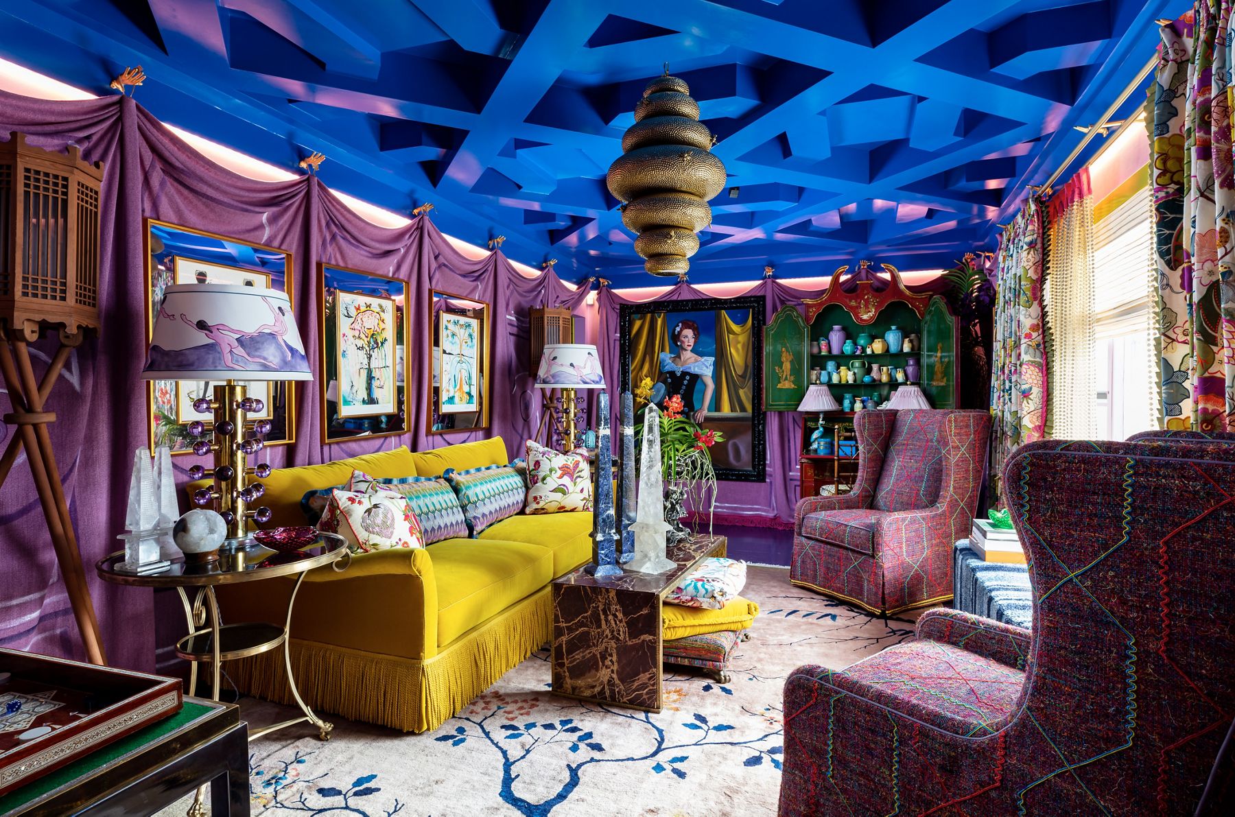 Un salon coloré comprend un canapé jaune, des fauteuils à oreilles à motifs rouges et sarcelles, une table basse en marbre, des tentures violettes et un plafond avec des conduits explosés peints en bleu vif.