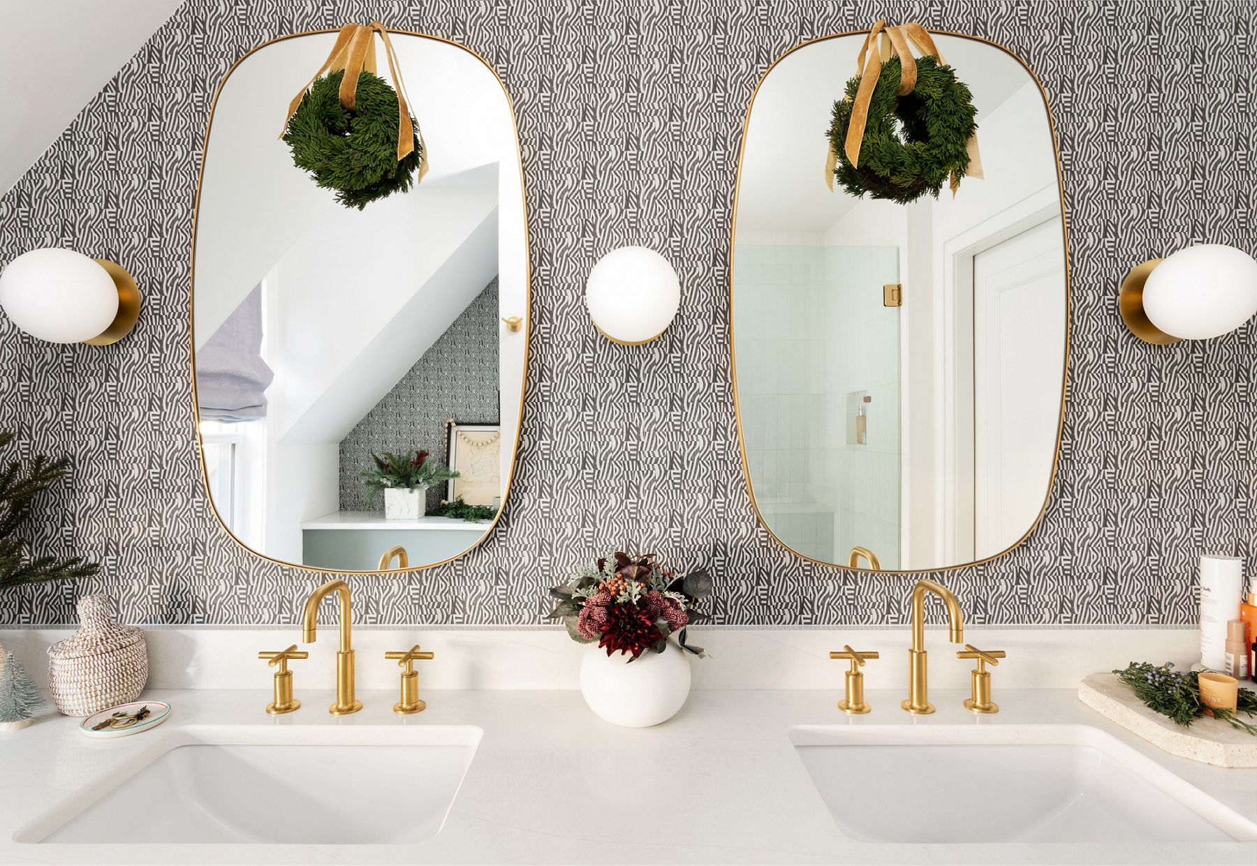 Centered in the image is a double-sink vanity space with two matching and symmetrical mirrors, under-mount sinks, brass faucets, and sconce lighting. A bold pattern wallpaper covers the wall and the mirrors and counter top are decorated with gold ribbon and green wreaths.