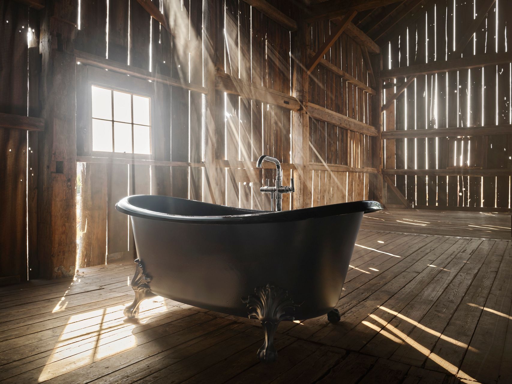 Une baignoire autoportante en fonte émaillée mise en valeur par des rayons de soleil spectaculaires qui pénètrent par la fenêtre d’une grange, créant un jeu saisissant d’ombres et de lumières sur le plancher.