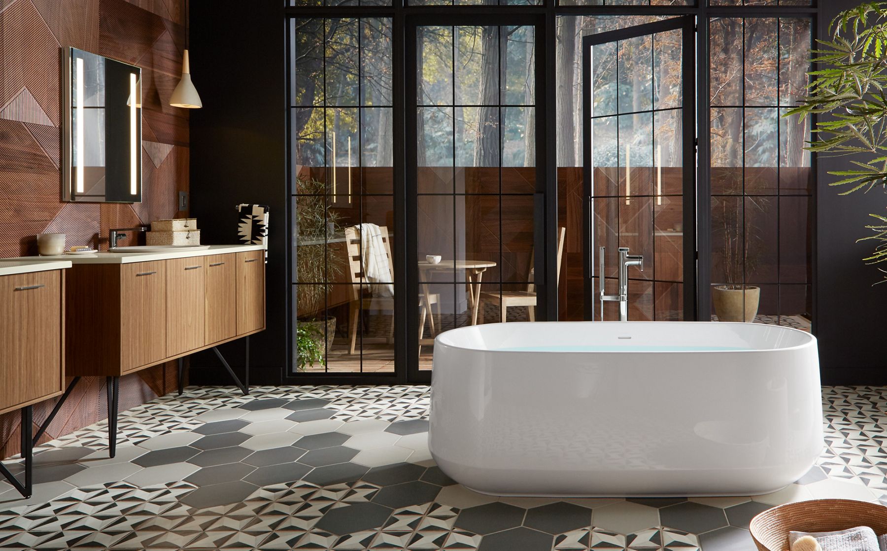 Une baignoire autoportante blanche Ceric de KOHLER avec remplisseur de baignoire à installation au sol Composed se tient devant des portes vitrées donnant sur un balcon avec une table et des chaises dans une salle de bain dotée de deux meubles-lavabos Jute sur pieds en épingle.