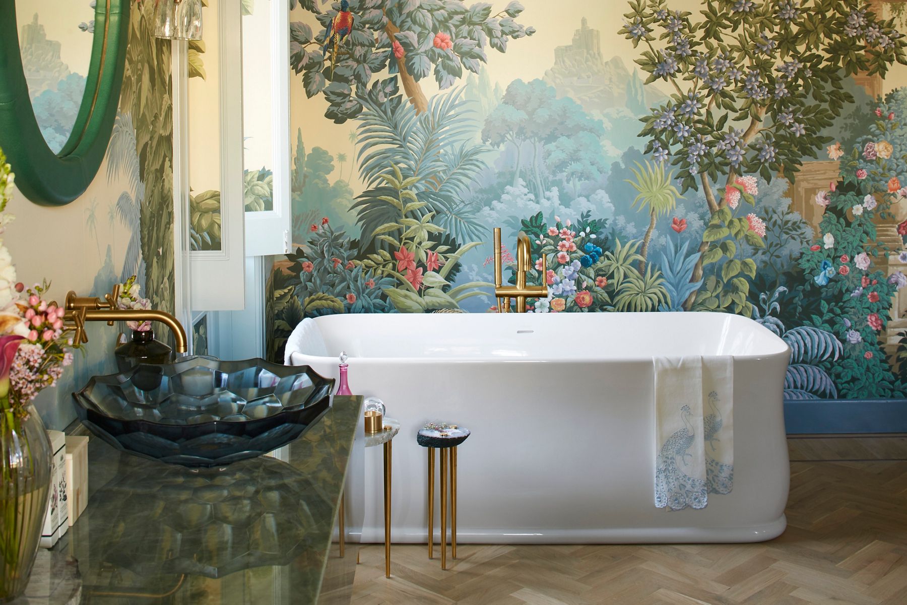 Un lavabo Briolette en verre bleu à facettes dans une salle de bain avec un papier peint jungle audacieux, une baignoire autoportante blanche, un robinet en laiton brossé moderne à fixation murale et un comptoir en pierre verte.