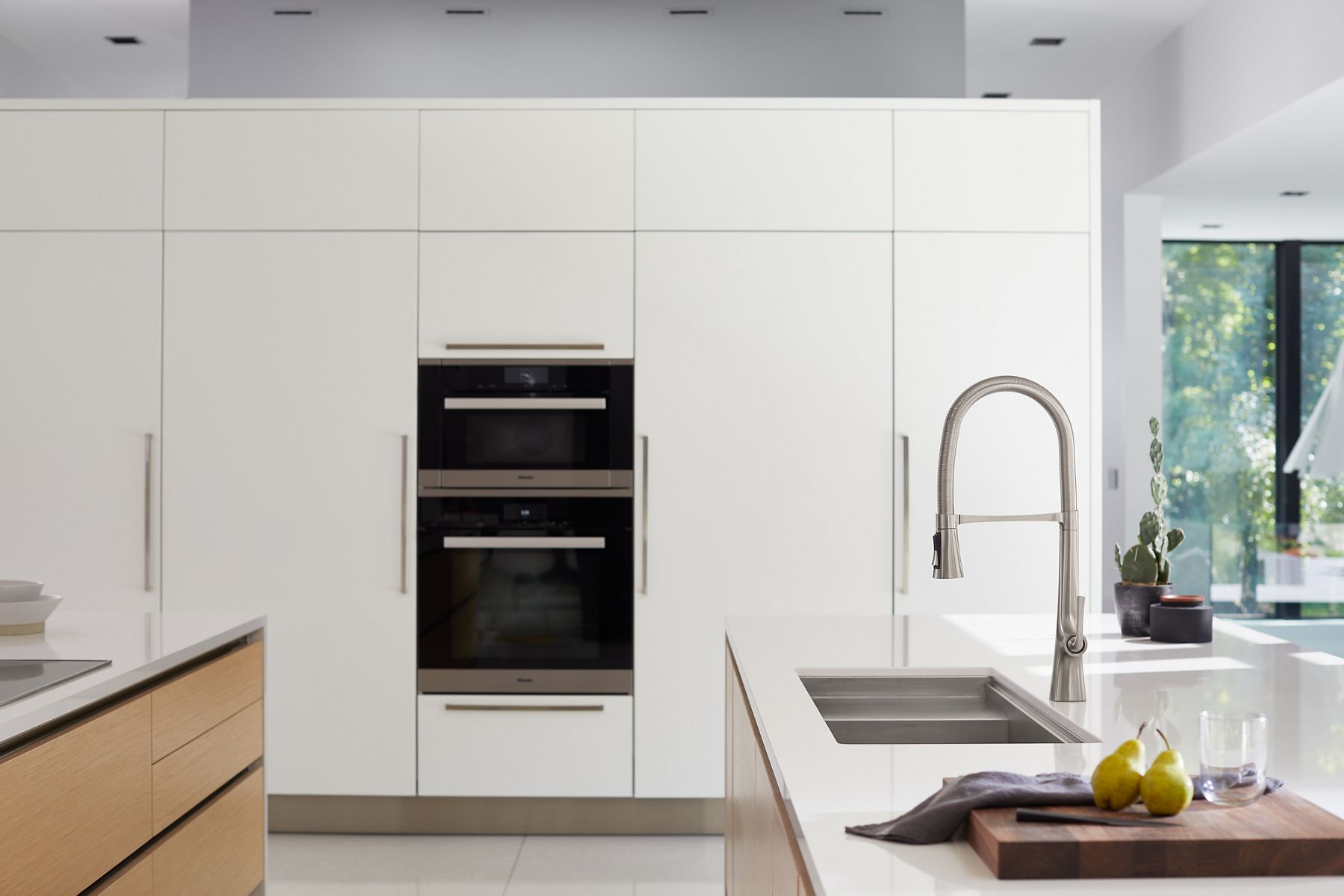 A sun-filled kitchen with white cabinets, a white counter top, and a stainless steel semi-professional faucet and stainless steel workstation sink. 