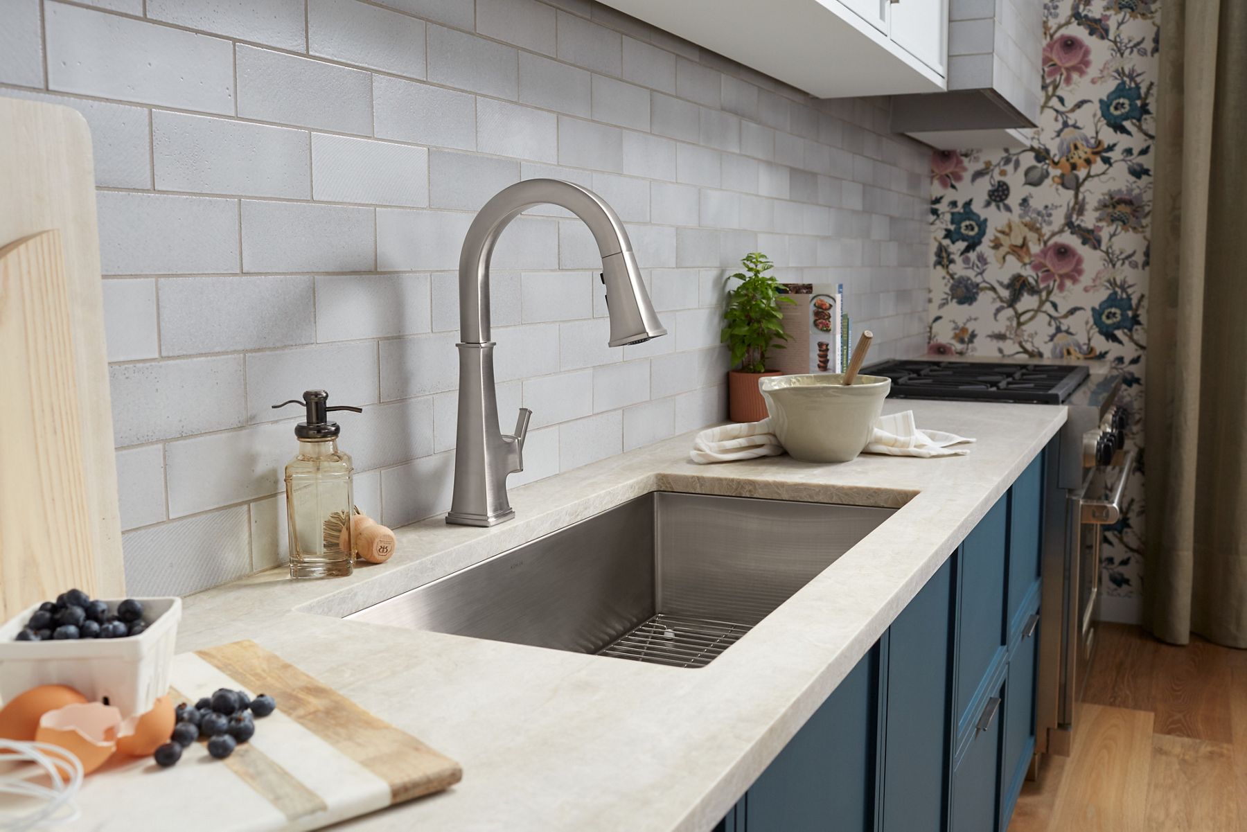 A kitchen with blue lower cabinets, white countertop, KOHLER Riff single-hole kitchen faucet with pull-out sprayer, white tile backsplash, and blue and white floral wallpaper.