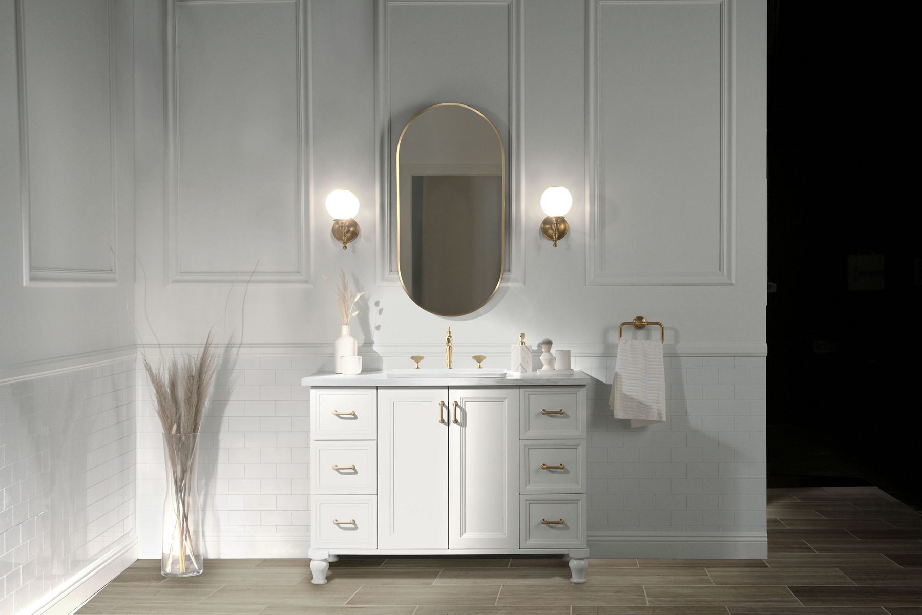 A bahroom styling space with a white vanity, pill-shaped mirror, and sconce lighting with brushed brass finishes.