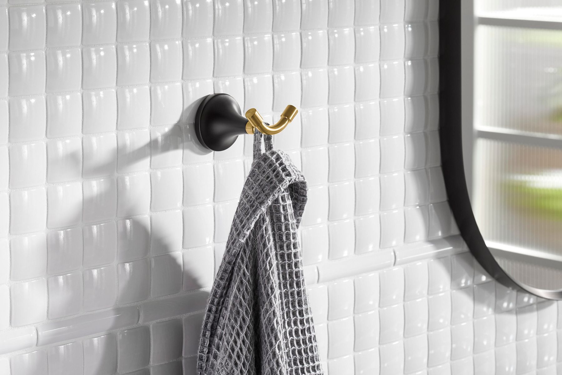 A gray robe hanging from a two-tone brushed brass and matte black robe hook attached to a glossy white tile wall.