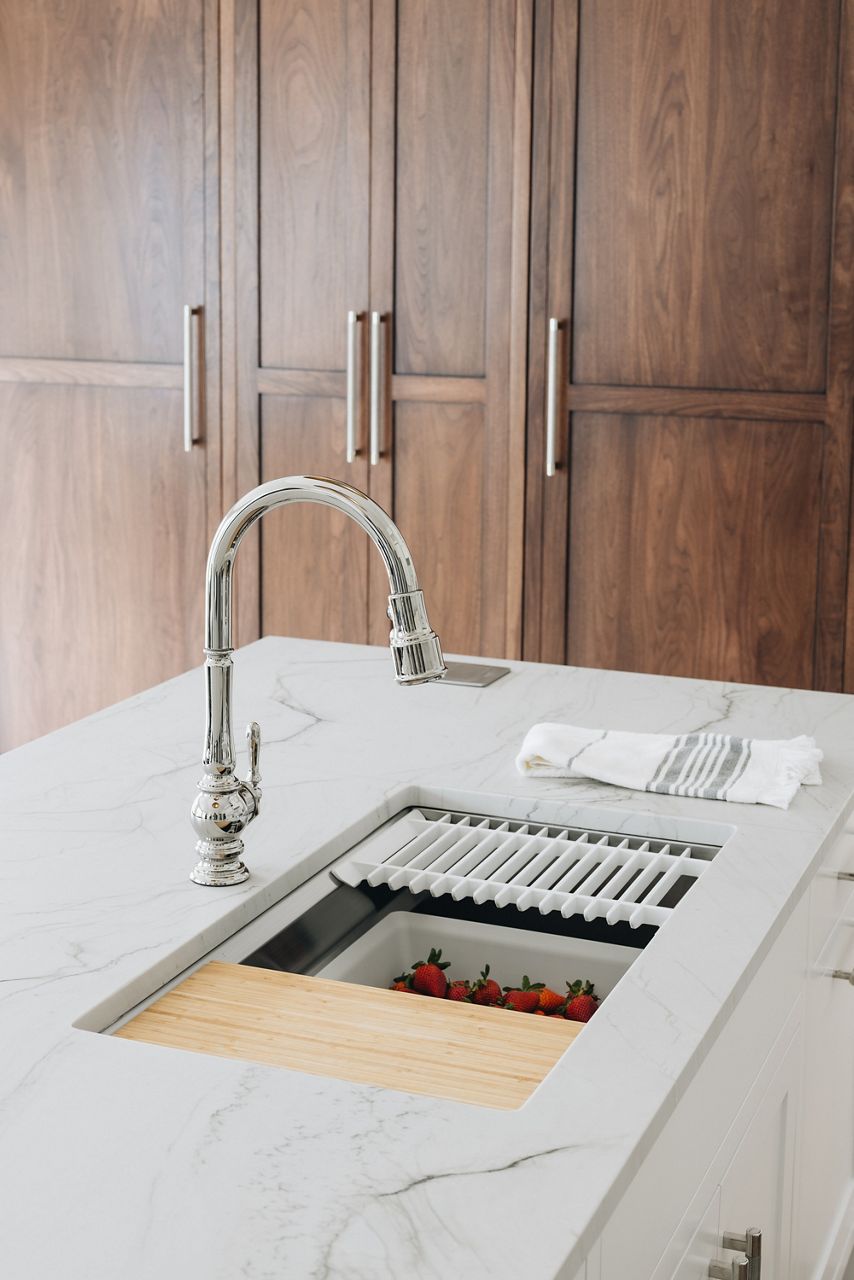 A traditional faucet workstation sink with various accessories in a kitchen island with a natural stone counter top.