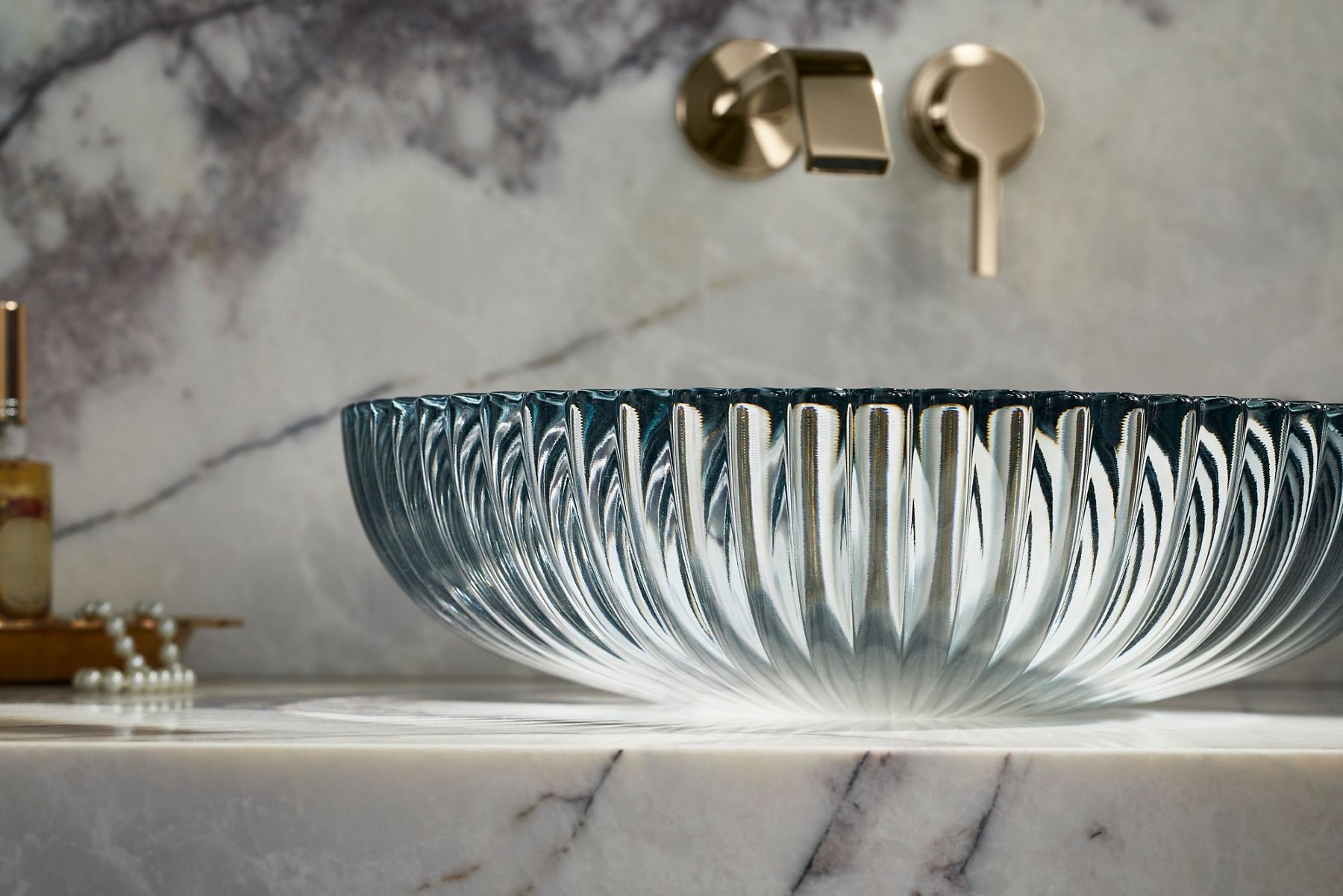Close-up of a round, fluted glass vessel sink with a radiating textured design, paired with a wall-mounted faucet on a marble vanity.