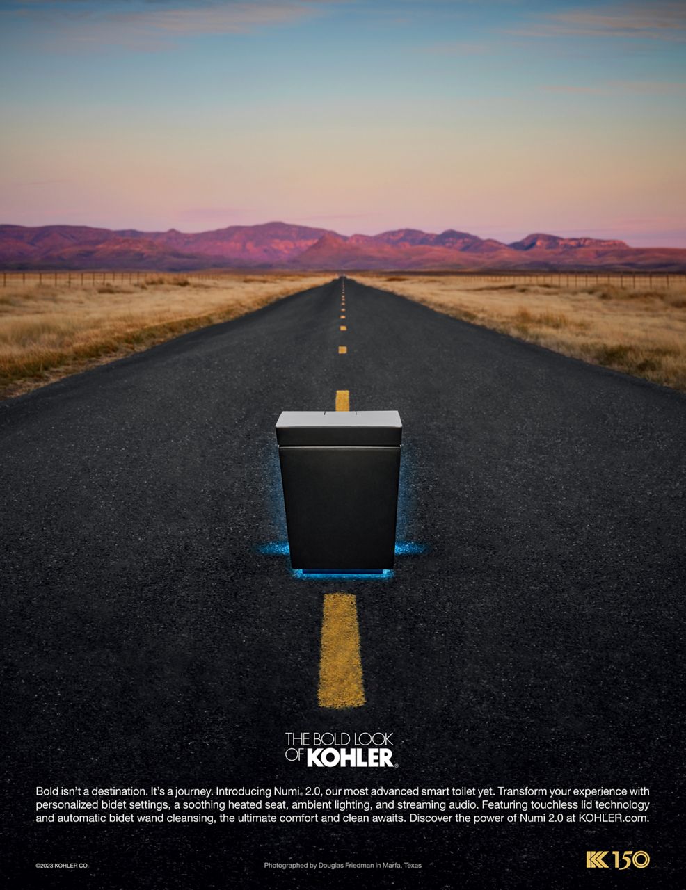 Image of the print advertisement featuring the matte black Numi 2.0 smart toilet in the middle of a long stretch of road, leading out to mountains reflecting a vibrant purple and pink sunset. 