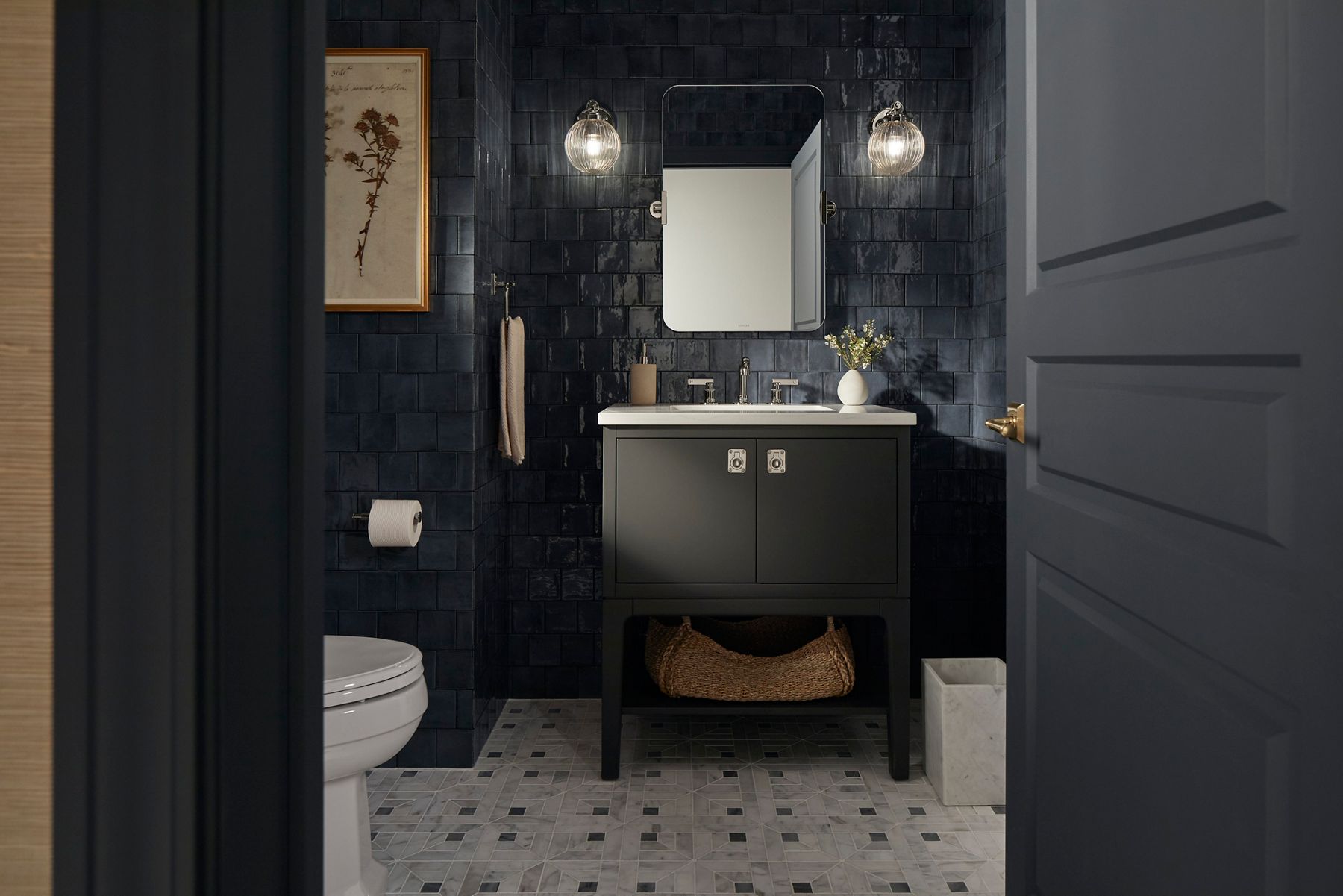 A dark gray vanity, chrome faucet, mirror, and sconce lights designed by Shea McGee in a bathroom with dark blue wall tile.