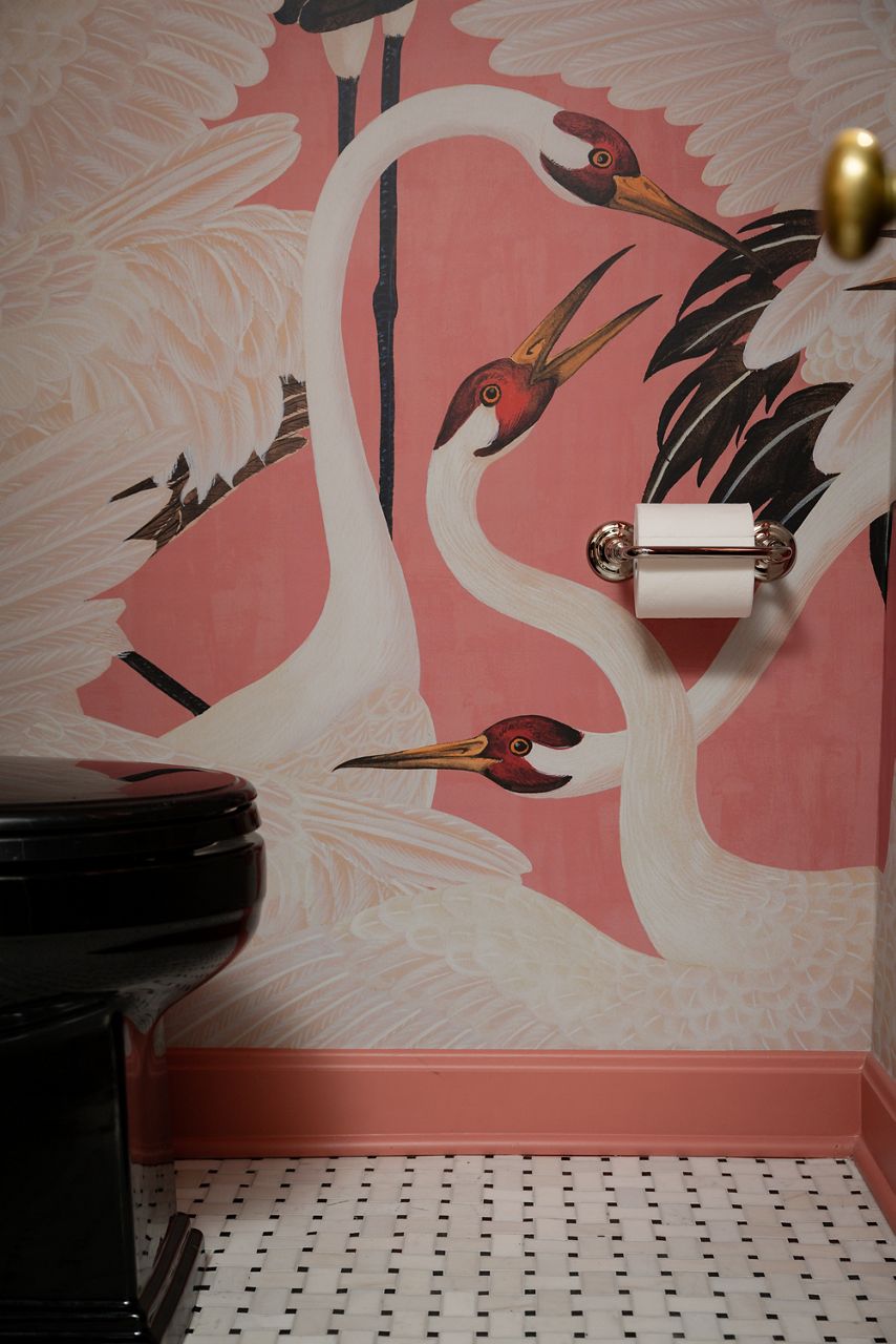 A close-up of a toilet paper holder with a roll of toilet paper against a pink wallpaper with a swan motif.