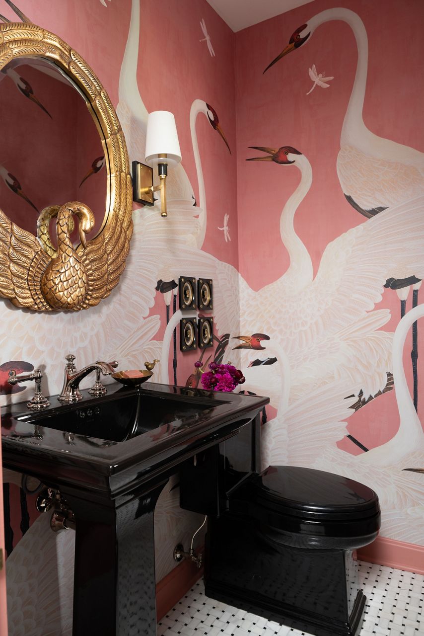 A stylish powder room with a black sink and toilet complemented by a pink wallpaper with a swan design and a vase with purple flowers on the counter.