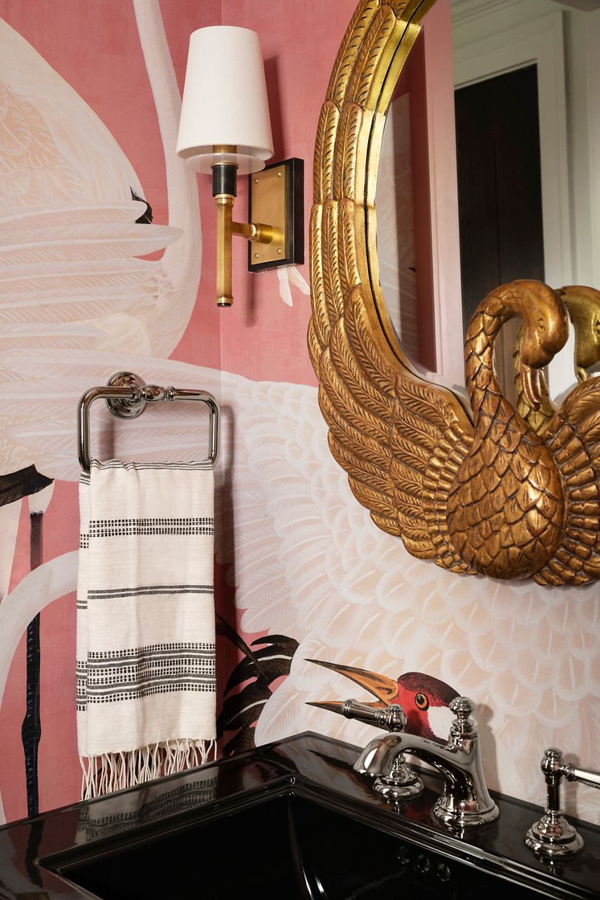 A close-up of a luxurious powder room featuring a gold mirror, black sink, and a striped towel on a metal towel ring set against a pink wallpaper with a swan motif.