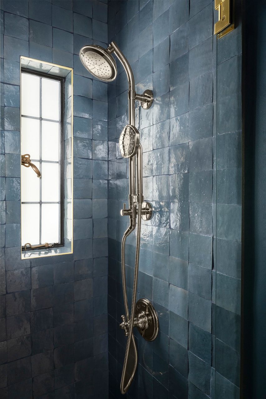 A close-up shot of a luxurious shower with dark blue tiled walls, brass shower fixtures included a showerhead and handshower sprayer, and a narrow frosted window with black framing.