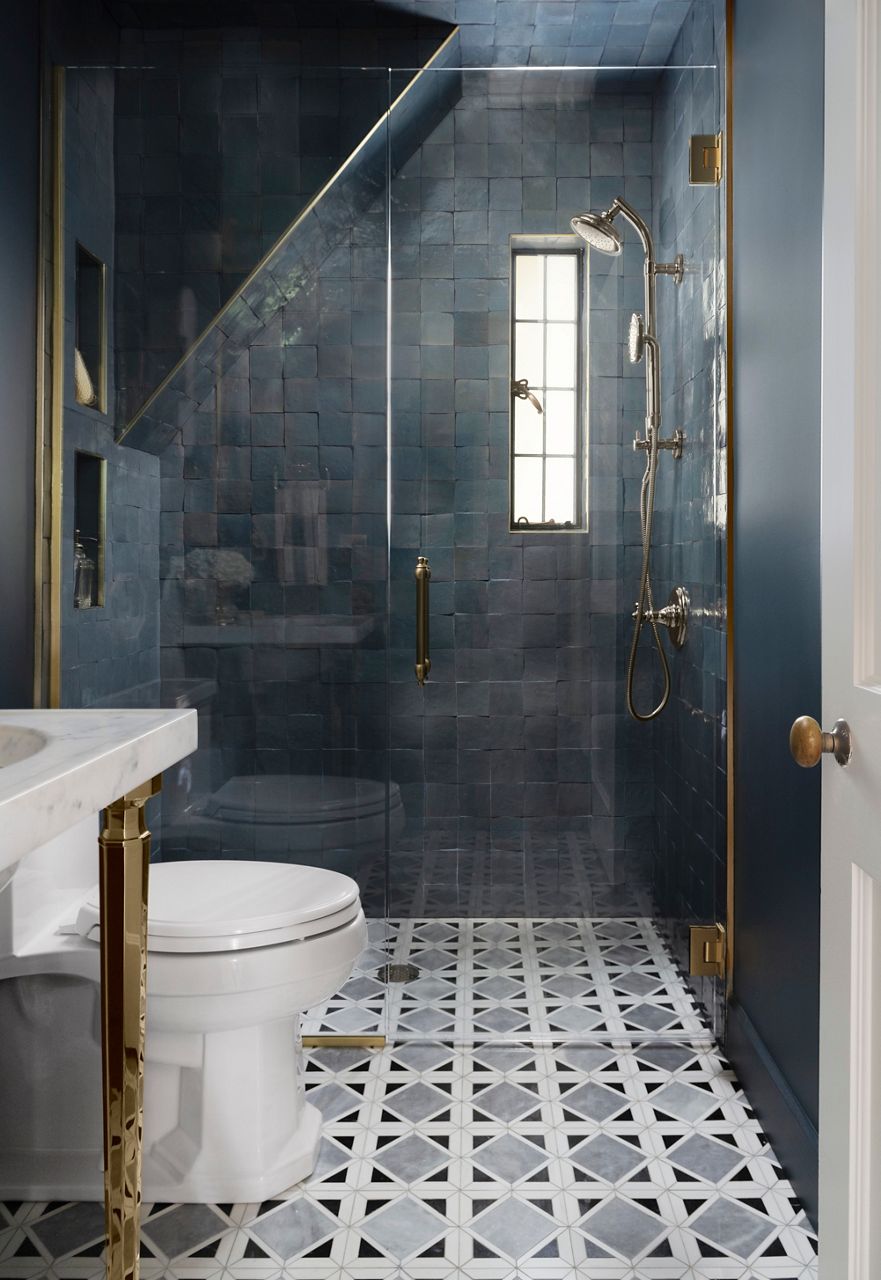 A portrait sunlit shot of a modern bathroom featuring a glass-enclosed shower with dark blue tiled walls, brass fixtures, a narrow window and geometric patterned floor tiles, adjacent to a white toilet and marble countertop.