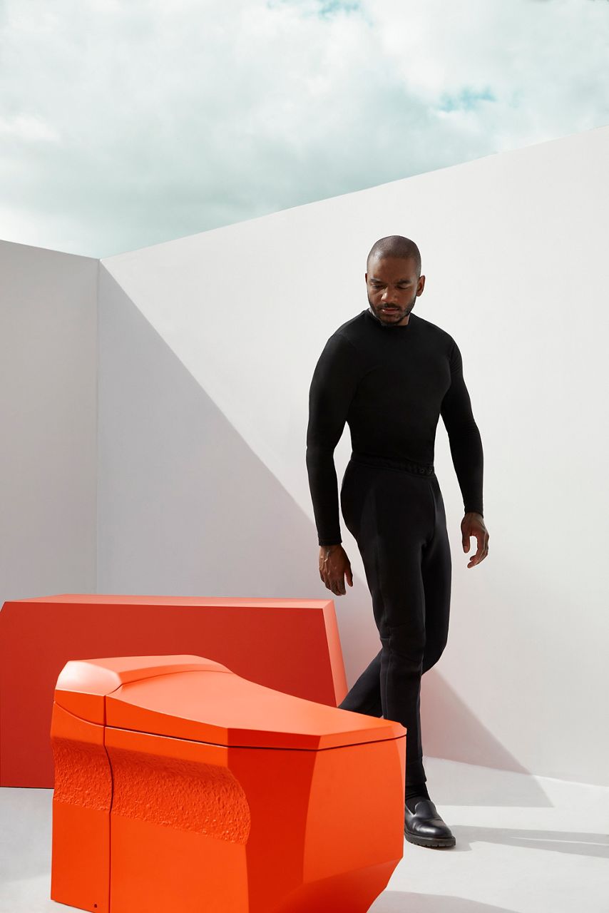 Artist and designer Samuel Ross looking at an orange toilet he designed in an outdoor room with white walls and no ceiling.