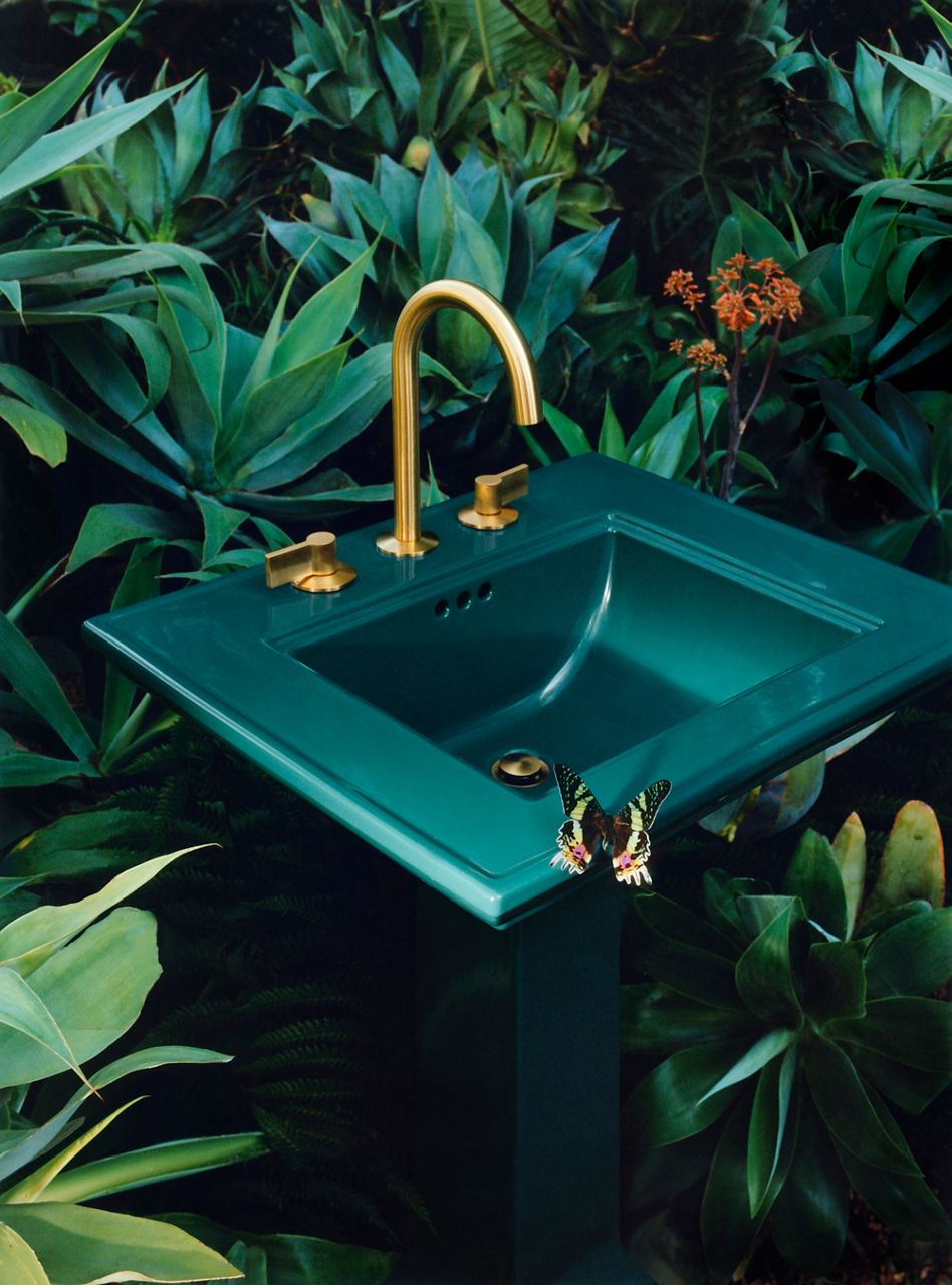 A portrait image of a teal standing bath sink positioned in the middle of a garden. The sink is designed with a gold faucet, gold handles, and a gold drainer. There is a yellow butterfly sitting at the edge of the sink.
