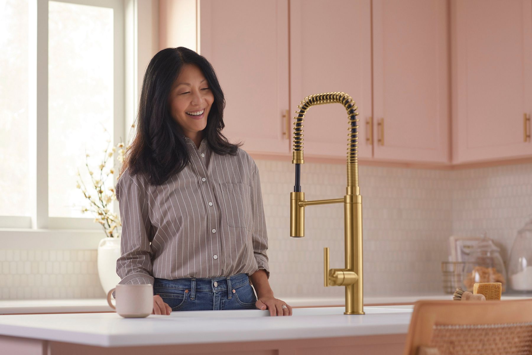A woman smiles while standing next to a modern gold kitchen faucet with a spring coil design in a bright kitchen. The background features light pink cabinets, a window with natural light, and kitchen decor.
