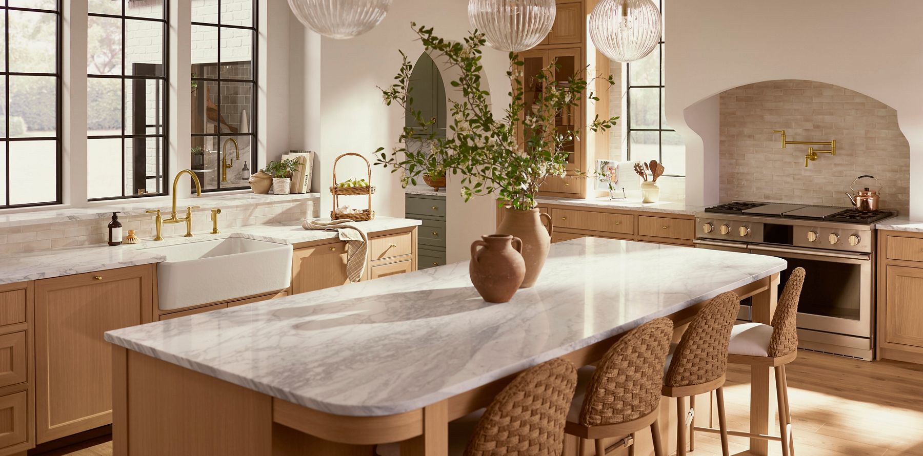 Bright kitchen featuring a large marble island, light wood cabinetry, open shelving, and decorative greenery.