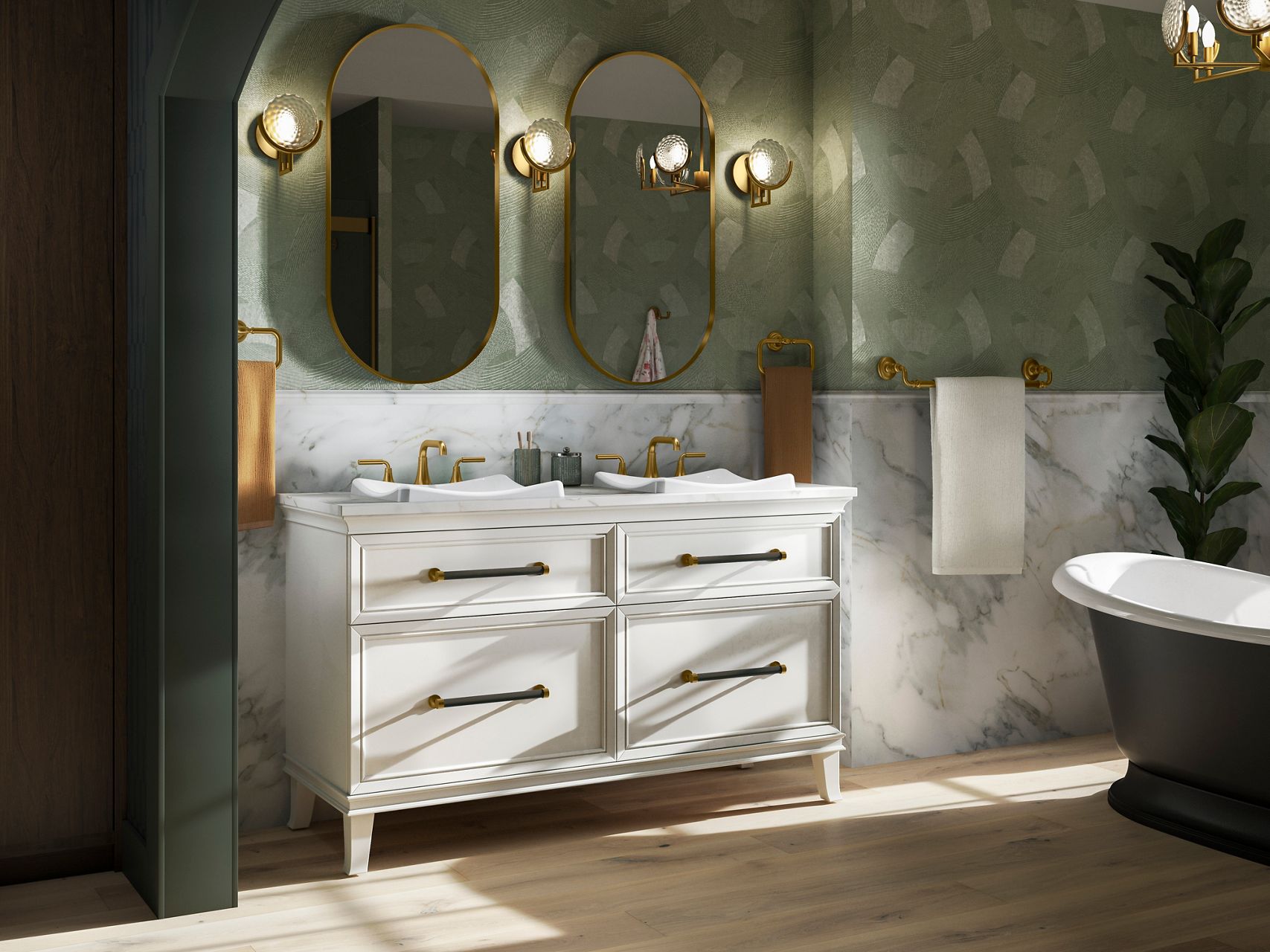 A landscape image of a double vanity area with two mirrors, two light scones, two sinks with gold faucets attached to a white cabinet storage unit. There is a tub on the righthand side that's matte black on the outside and white inside with a view of a towel on a towel rack and a fig leaf tree.