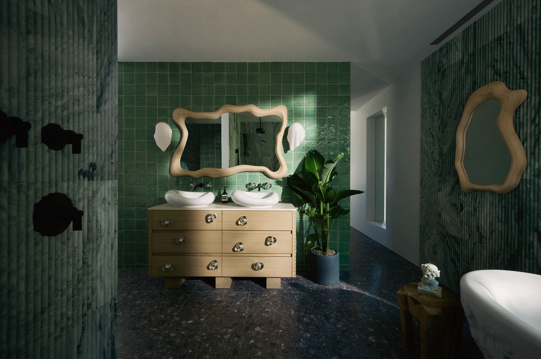 Modern bathroom with a double vanity featuring sculptural sinks, artistic mirrors with wavy frames, and green tiled walls. A large plant adds a touch of nature, and a small decorative bust is placed on a wooden side table near a textured marble wall.