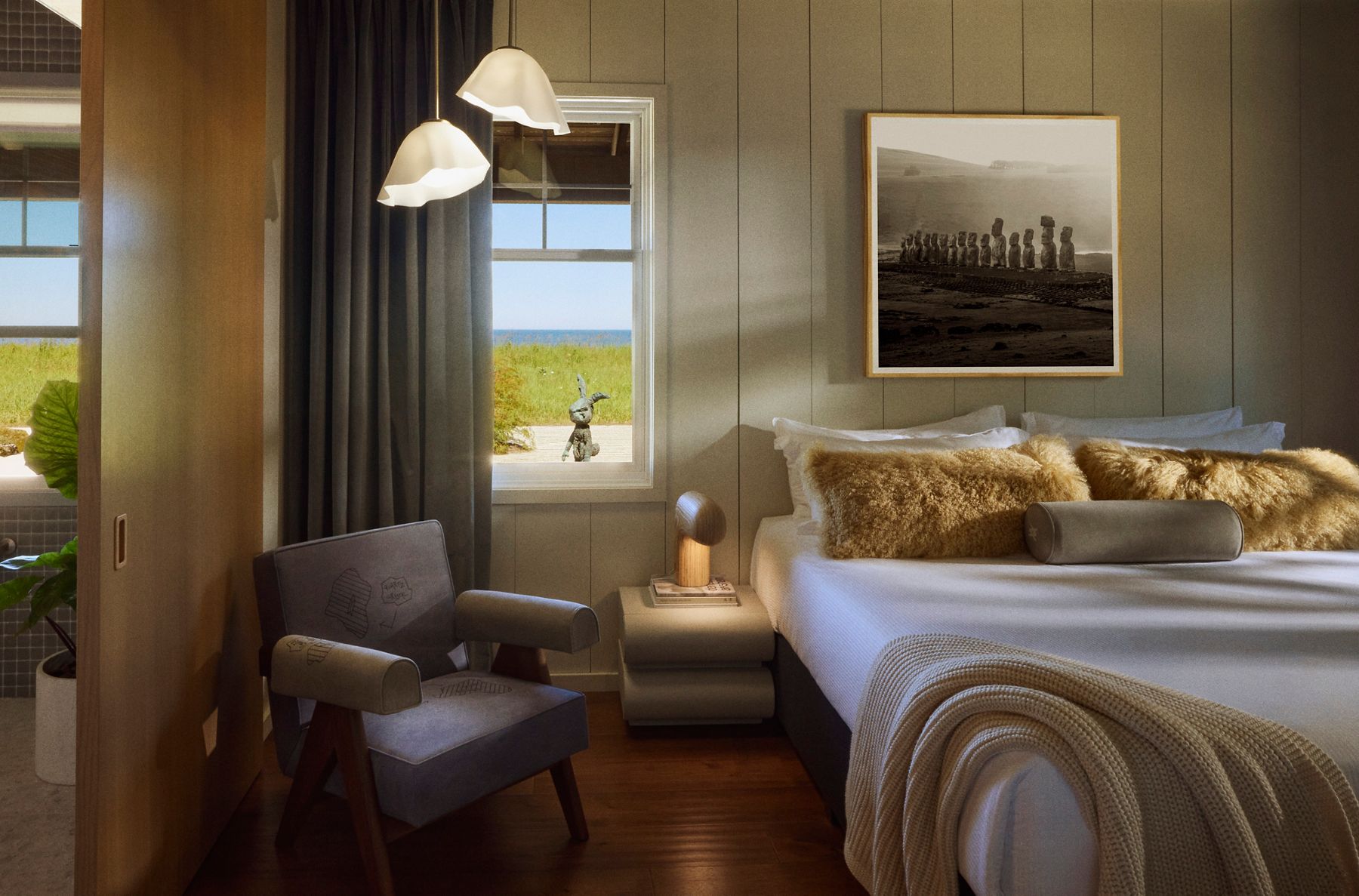 Cozy bedroom interior with a neutral color palette featuring a bed with textured pillows, a side table with a sculptural lamp, a unique chair, and a large framed photograph on the wall. A window reveals an outdoor view of greenery and the ocean.