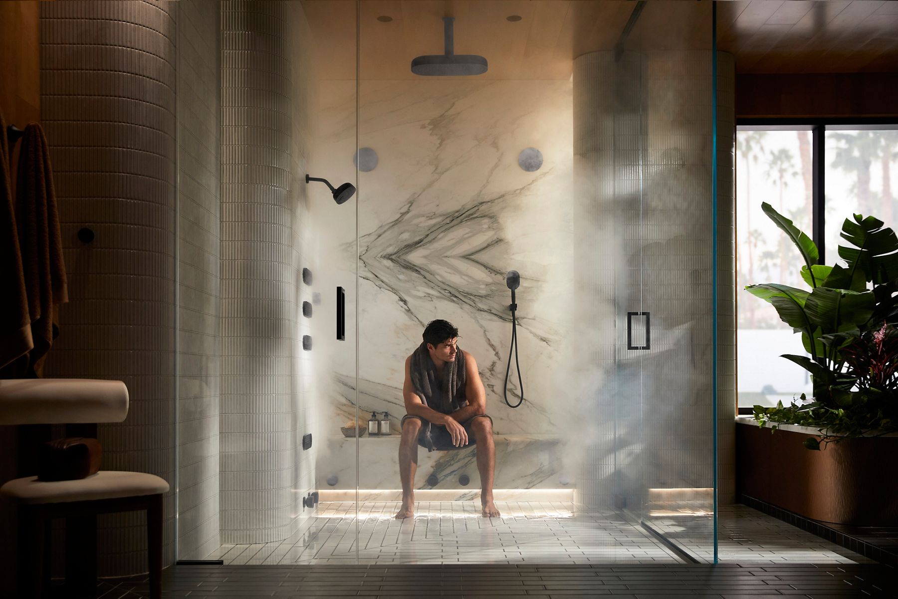 Man with towel sitting on a shower bench in a modern shower space with steam standing under water spray in a large shower while pressing digital remote with Matte Black showering components mounted to wall.