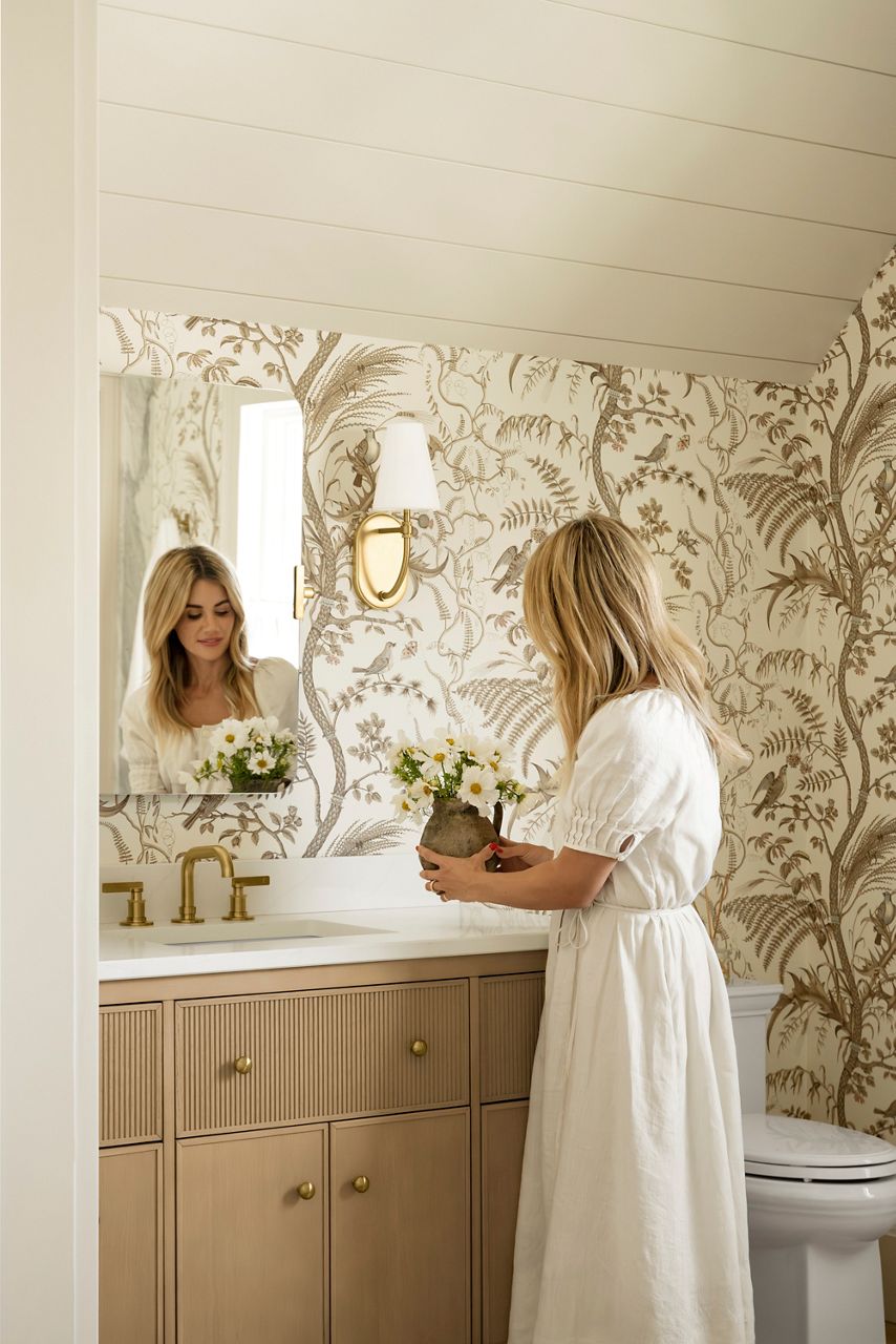 a woman in a white dress stands in a stylish bathroom featuring a vanity with brass fixtures, a white counter top, and floral-patterned wallpaper.