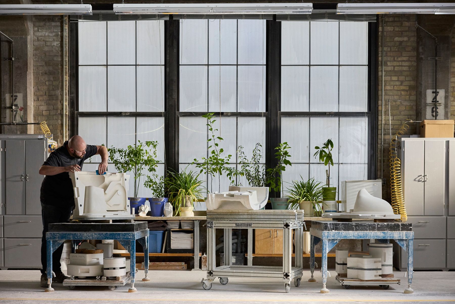 Artisan working at a long table in a bright industrial studio, assembling or refining ceramic pieces with tools, with multiple workstations and large grid windows in the background.