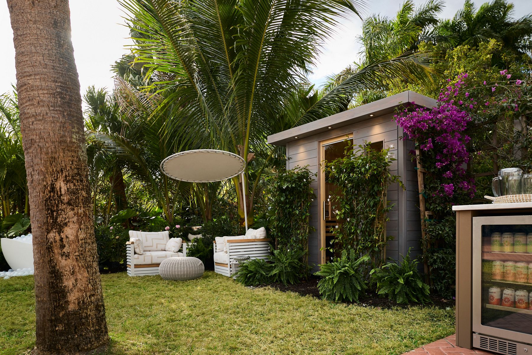 Outdoor sauna set in a tropical landscape and garden with cushioned lawn chairs and umbrella.