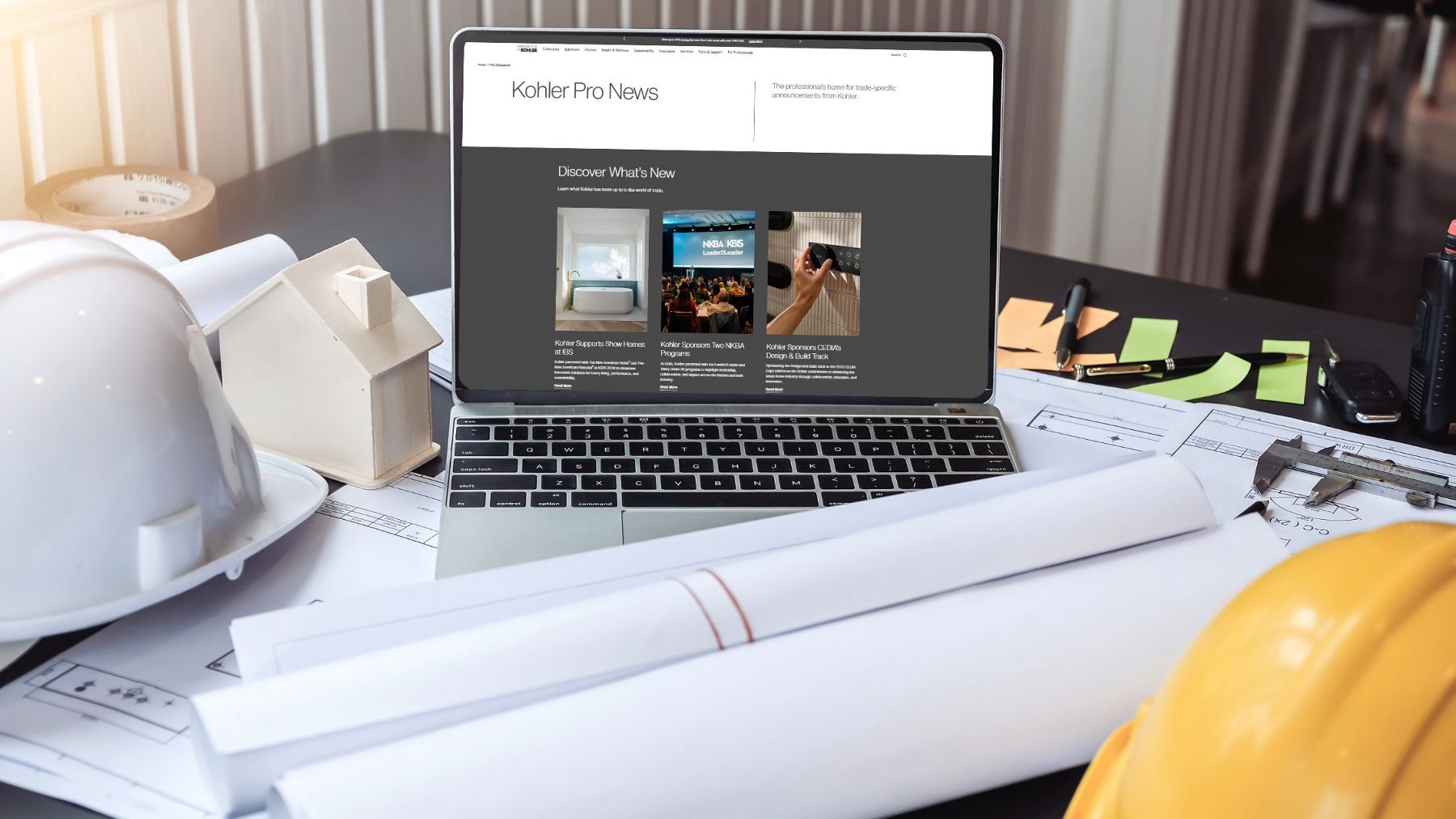 Laptop with an open browser on a table, next to yellow and white contrusction hats and paperwork with blueprints and rolled up files.