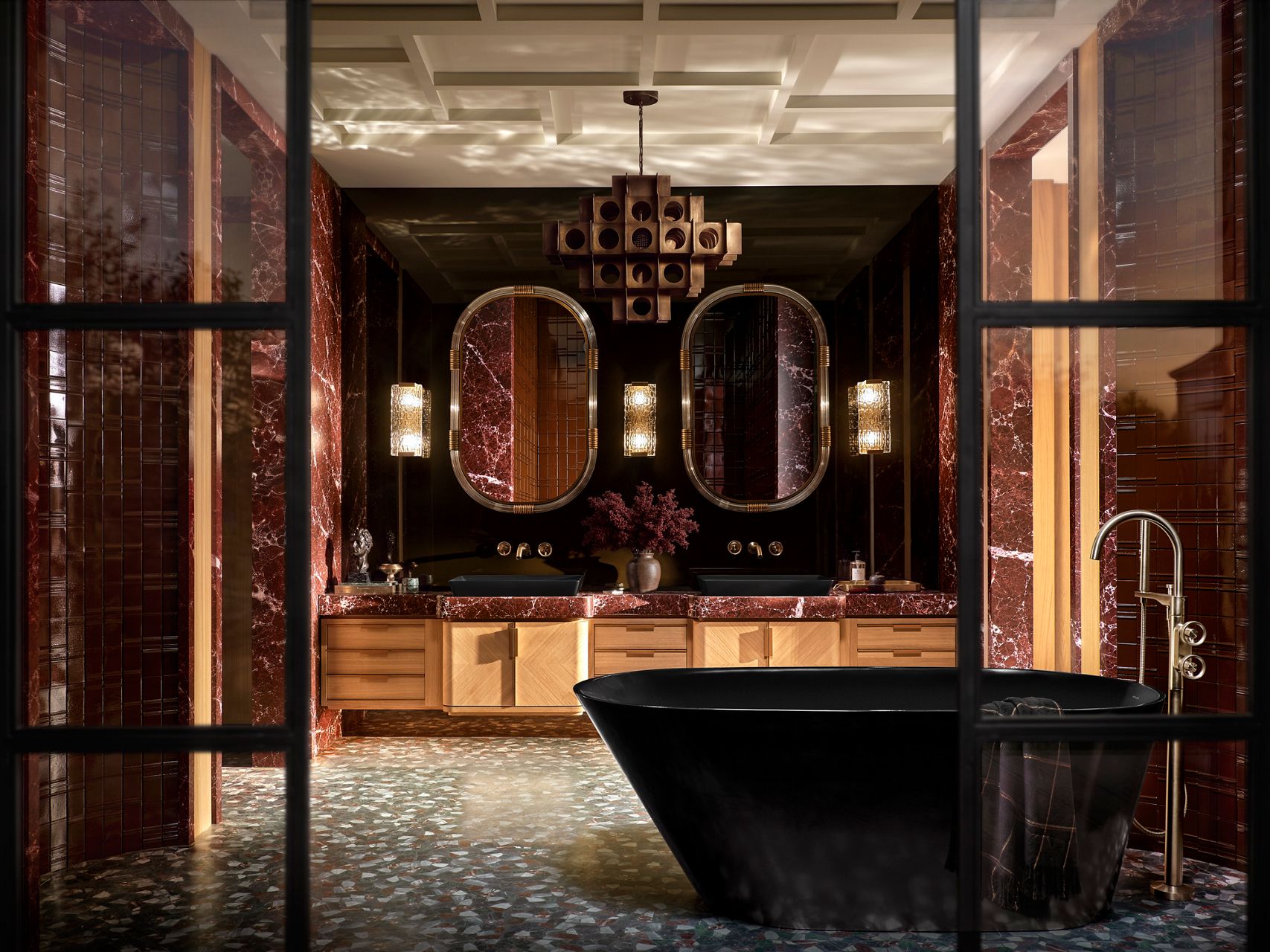 Dramatic bathroom with a black freestanding bathtub, red marble walls and countertops, double vanity, and warm wood cabinetry.