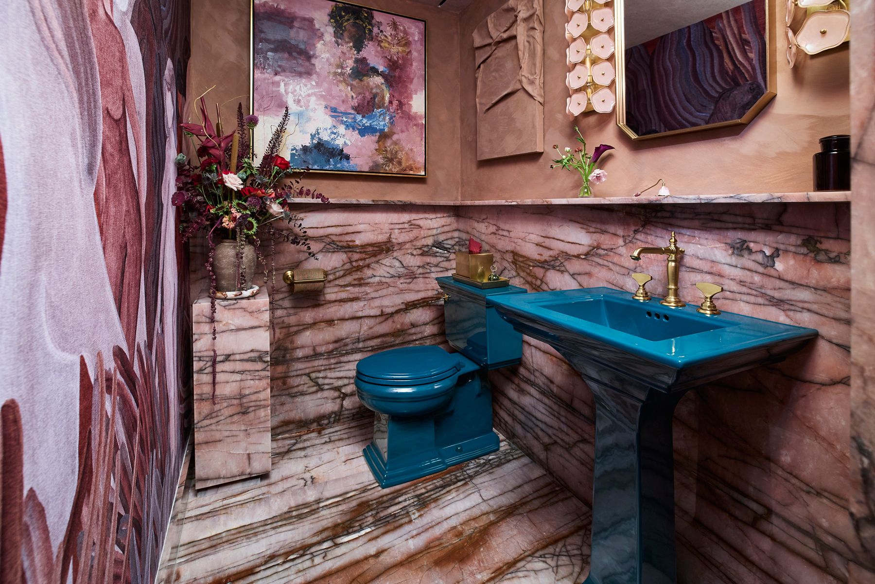 Dramatic powder room featuring pink veined marble walls and flooring, a deep blue pedestal sink and toilet, and polished brass fixtures.