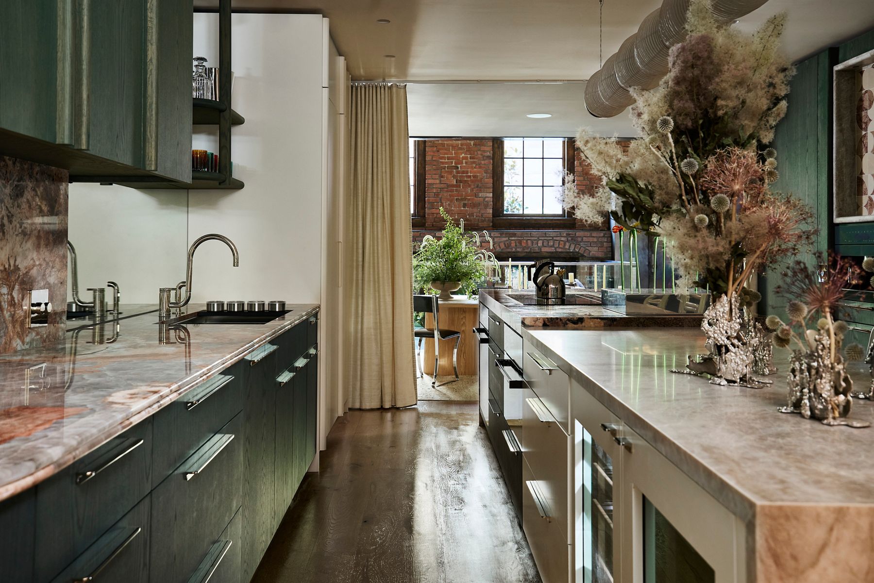 Modern kitchen with dark green cabinetry, expansive stone countertops, stainless steel faucets, and floral arrangements overlooking an exposed brick living space.