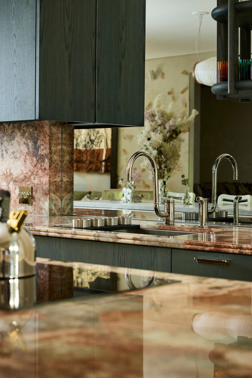 Close-up of a polished stainless steel kitchen faucet and sink set into richly veined stone countertops with dark green cabinetry.
