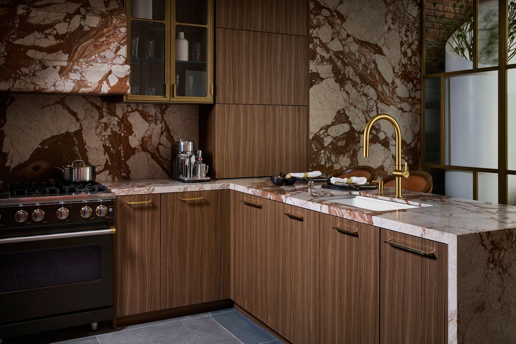 Sophisticated kitchen featuring warm wood cabinetry, dramatic red and cream veined stone surfaces, and a brushed brass faucet over a white undermount sink.