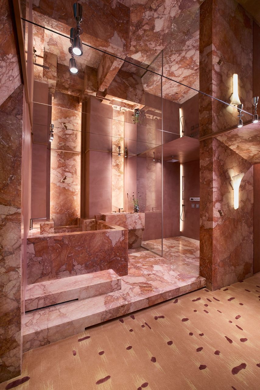 Luxurious bathroom clad in rose-colored marble with a glass-enclosed shower, integrated stone bathtub, mirrored walls, and warm ambient lighting.