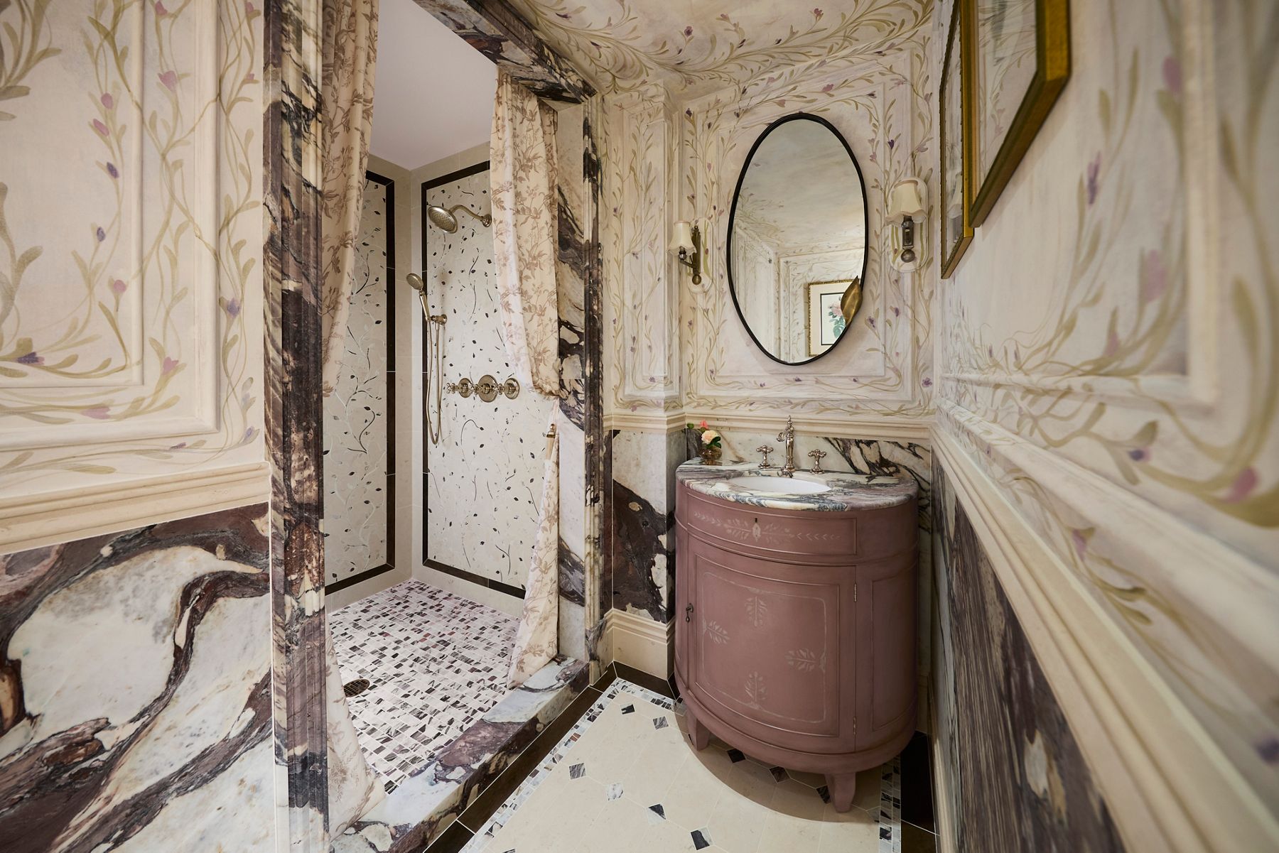 Ornate bathroom featuring hand-painted floral walls, richly veined stone accents, a dusty rose vanity, oval mirror, and walk-in shower with brass fixtures.
