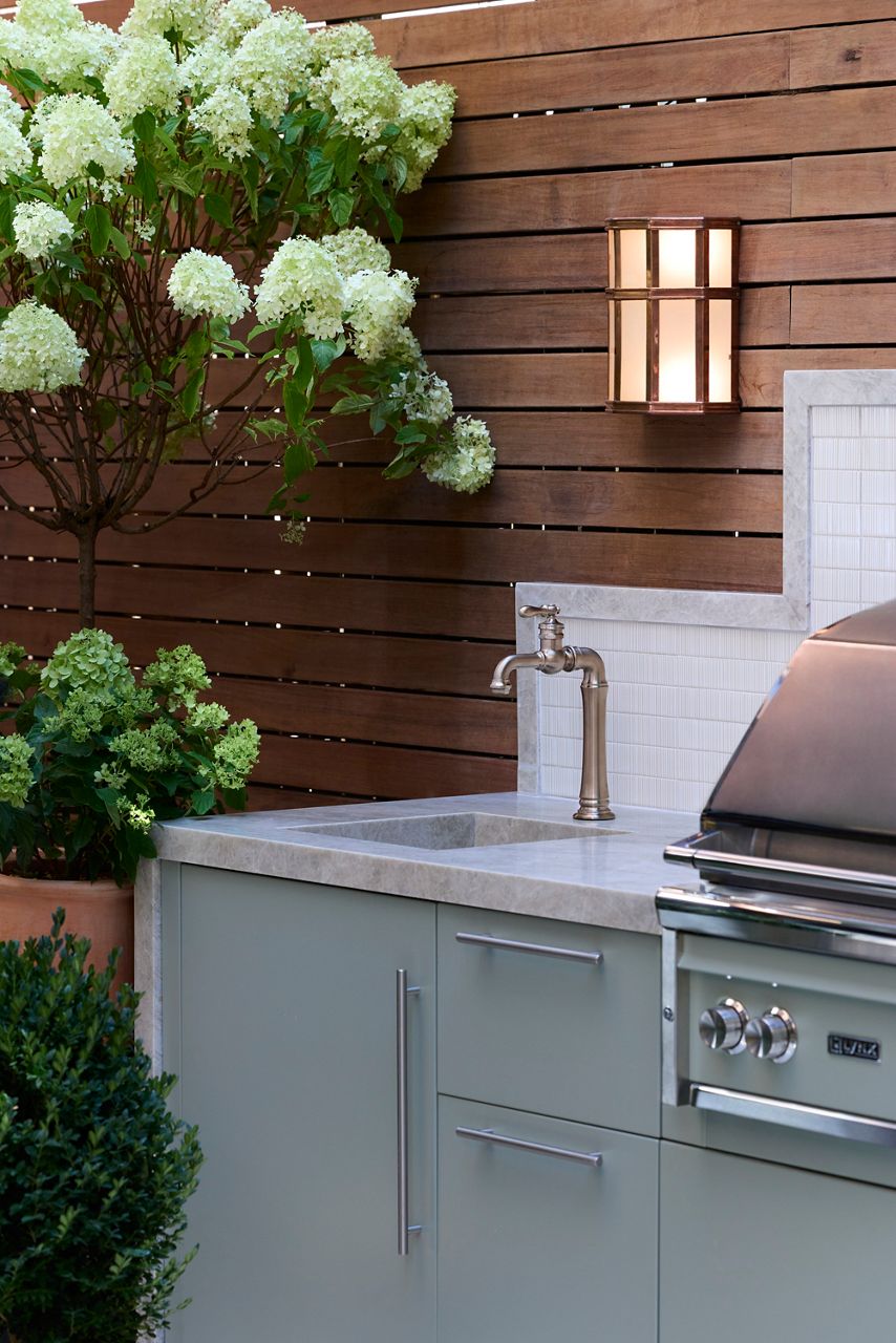 Outdoor kitchen with sage green cabinetry, stone countertop and sink, traditional metal faucet, built-in grill, wood privacy wall, and flowering hydrangeas.