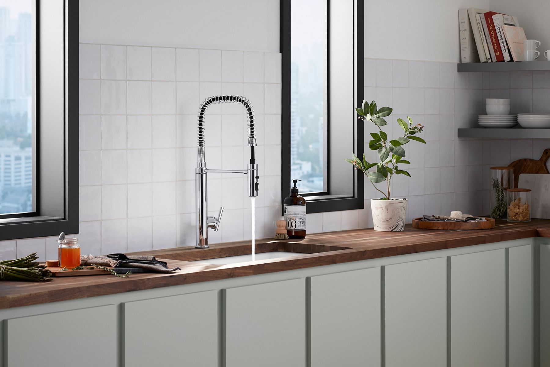 Modern kitchen sink workstation with a spring coil faucet, wood countertop, white tile backsplash, and black-framed windows.