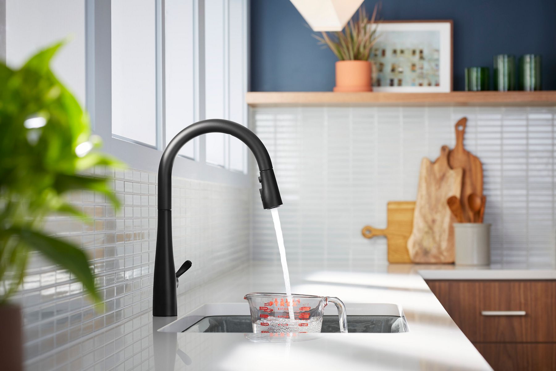 Matte black kitchen faucet dispensing water into a measuring cup at a white sink in a modern kitchen.
